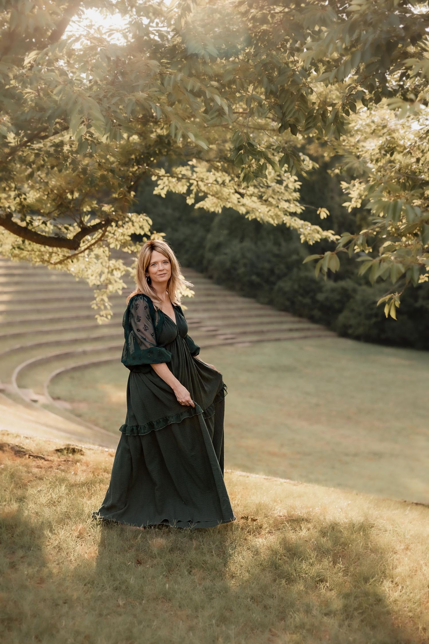 Woman in dark green gown standing in sunlit park with stone amphitheater steps and lush trees in background.
