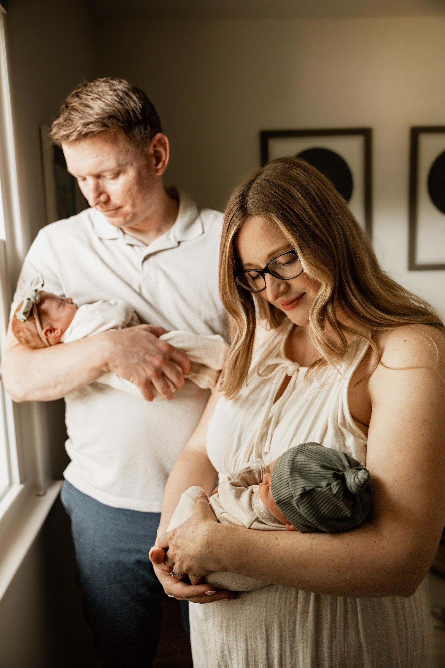 A couple tenderly holds their twin newborns near a bright window in a warm, intimate indoor newborn photography session.