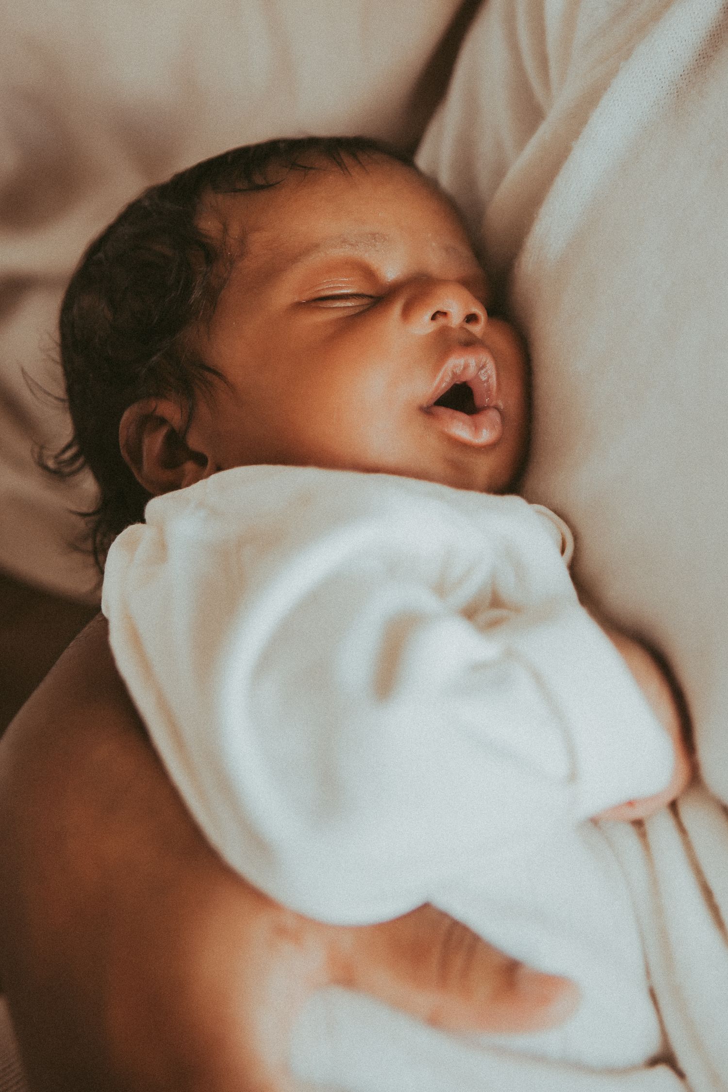 A sleeping infant swaddled in soft white fabric rests peacefully against light-colored bedding.