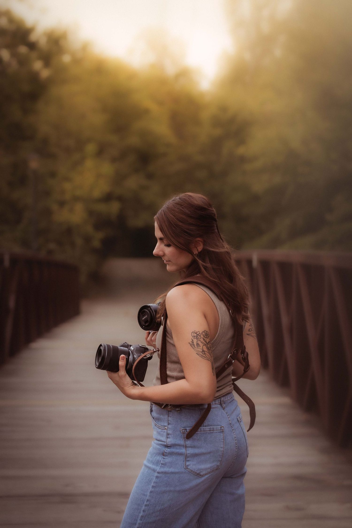 Photographer with long brown hair holding cameras on wooden bridge during golden hour sunset.
