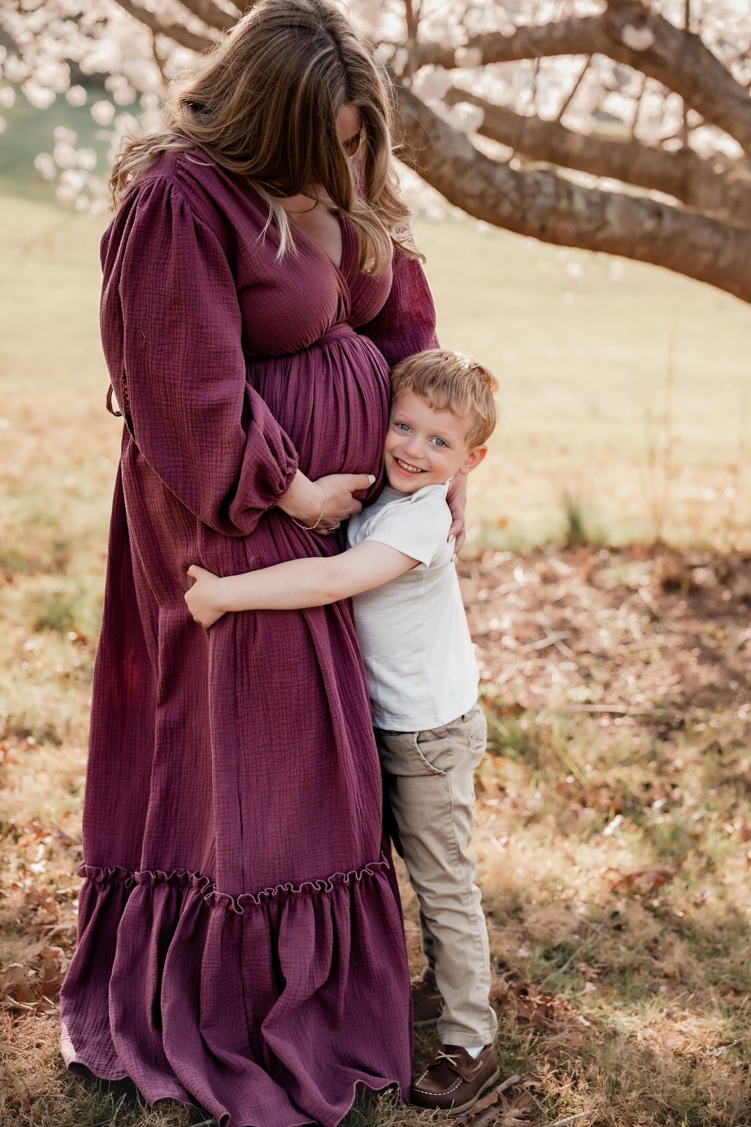 A young boy hugs his pregnant mother in a burgundy dress outdoors near a blossoming tree.