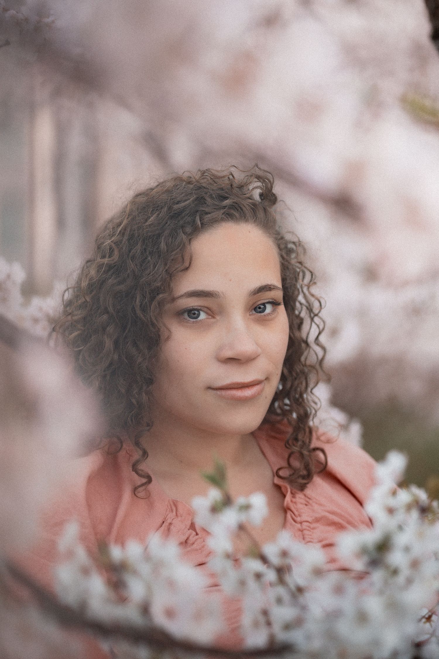 Young woman with curly hair in pink blouse surrounded by blooming cherry blossom branches in spring.
