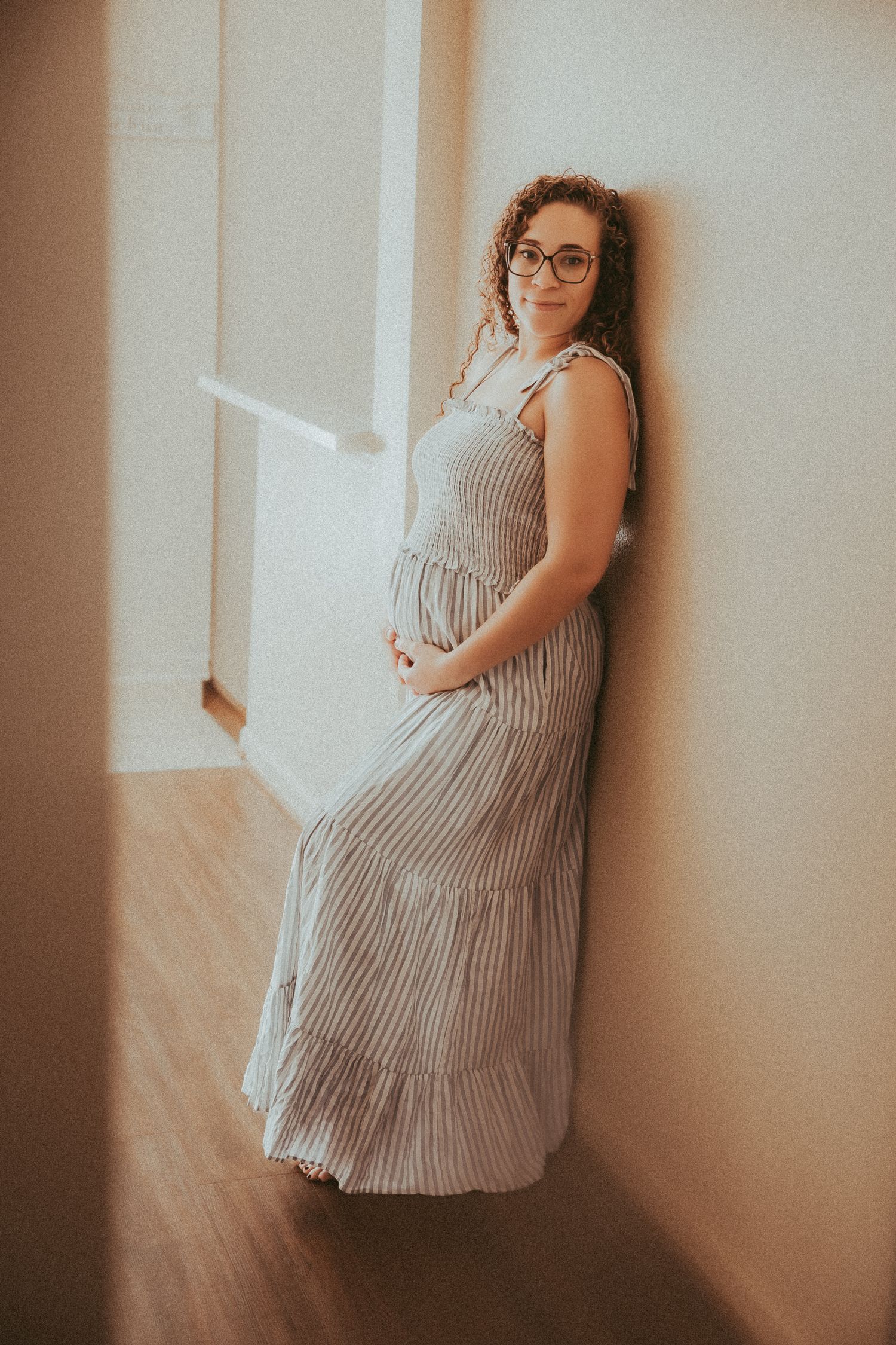 A person in glasses wearing a striped maxi dress poses against a wall in soft, warm lighting.