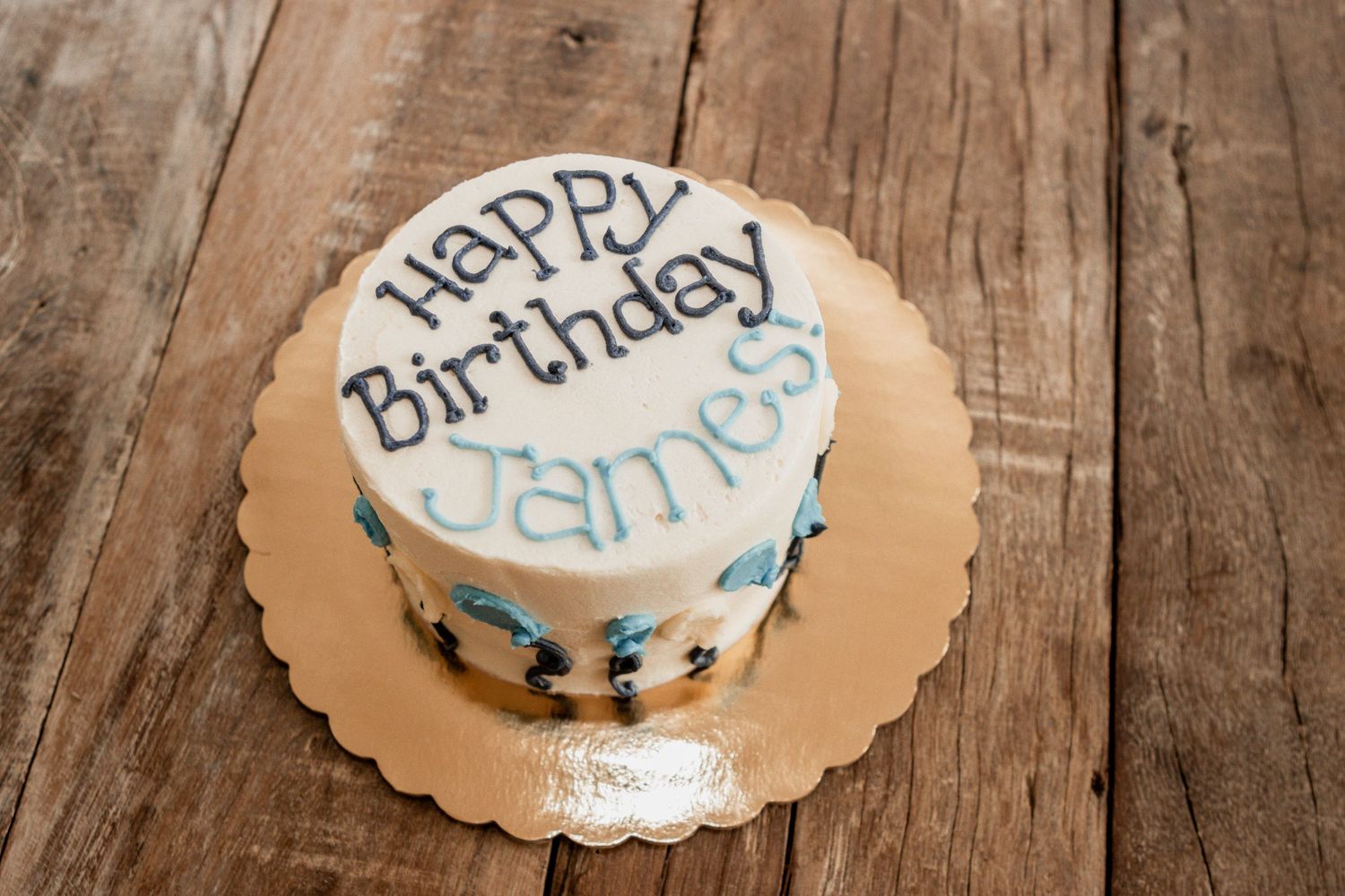 Three circular birthday cakes with white frosting and blue accents displayed on wooden table.