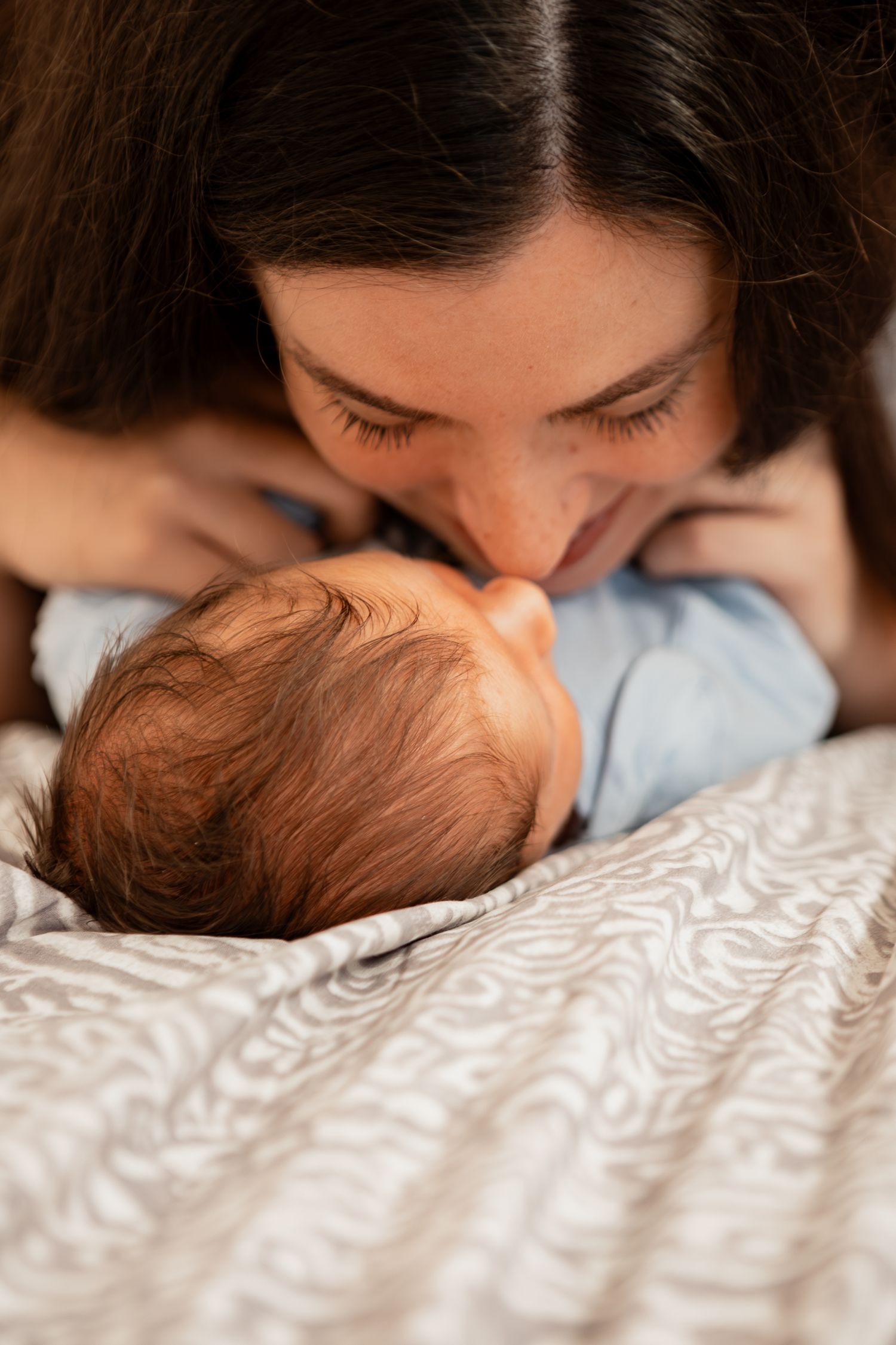 Baby with brown hair lies peacefully on white patterned bedding while being comforted.