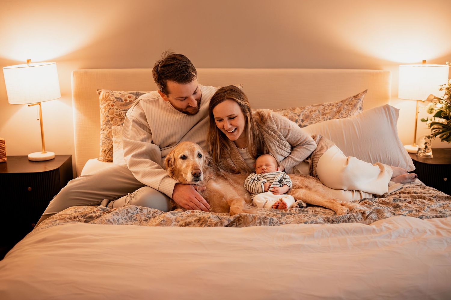 Couple relaxes on bed with golden retriever dog and baby in cozy bedroom with warm lighting.