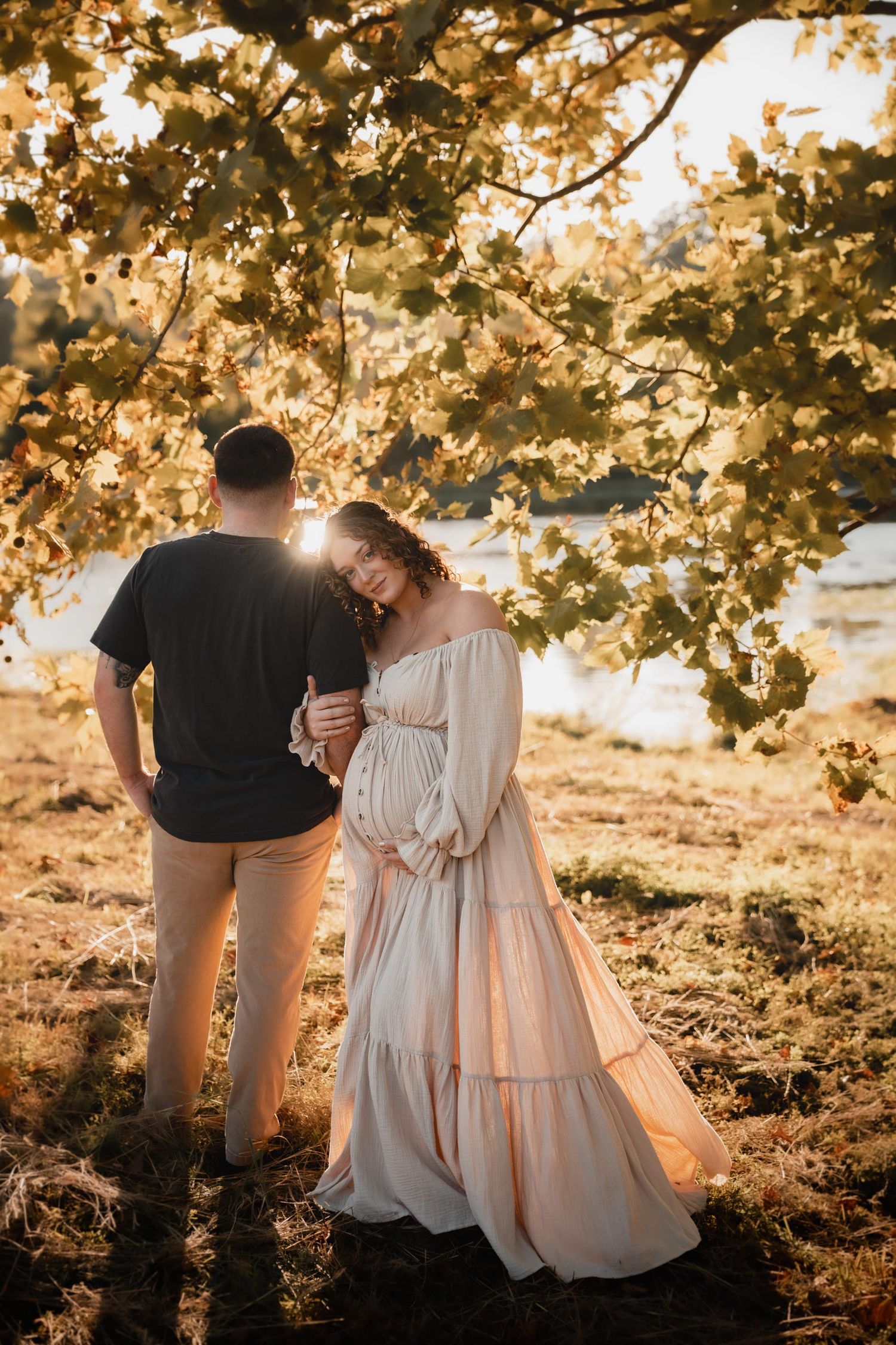 Pregnant couple embracing under golden autumn tree at sunset, woman in flowing white dress cradling baby bump.