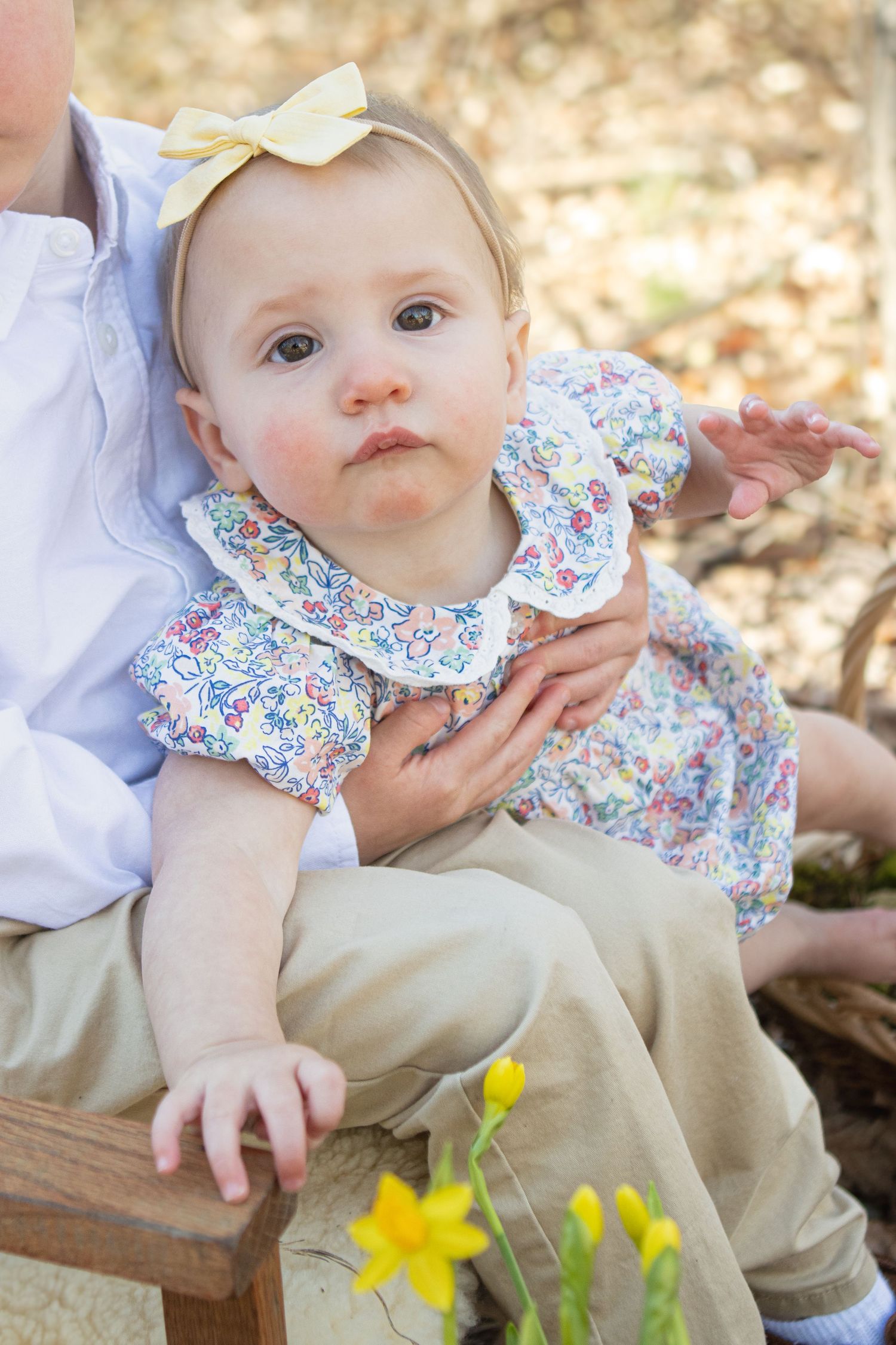 Baby in floral dress with bow sits on wooden chair with daffodils blooming nearby.