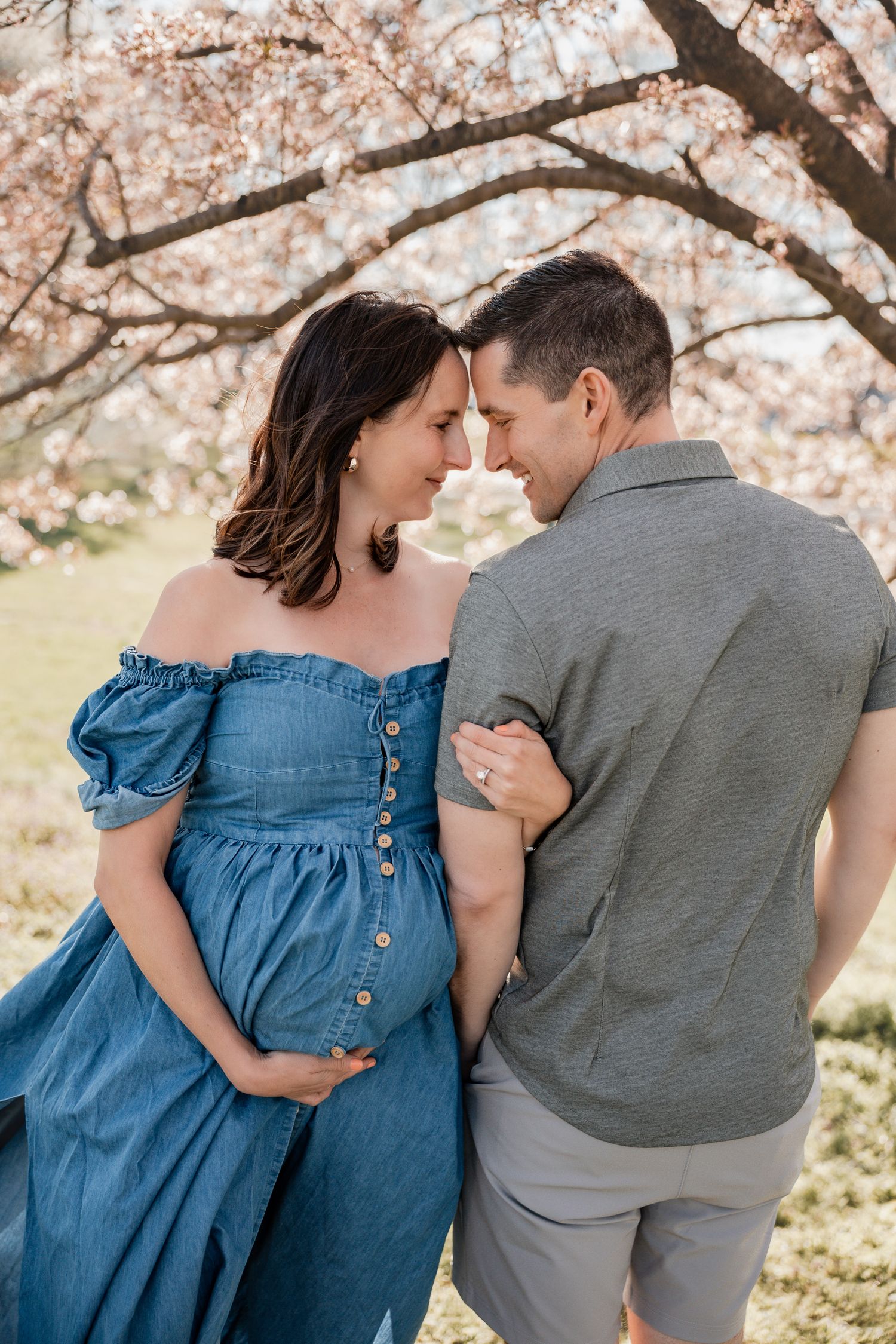 Pregnant couple sharing a tender moment under blooming cherry blossom trees, woman in blue off-shoulder dress.