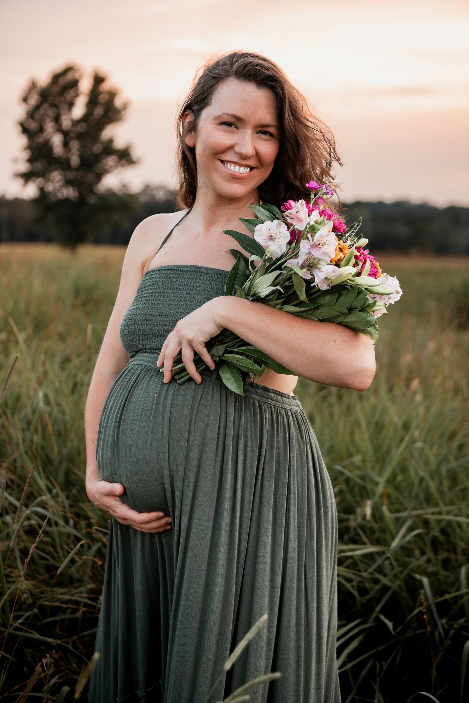 A woman in a sage green maxi dress holding fresh flowers in a golden sunset field.