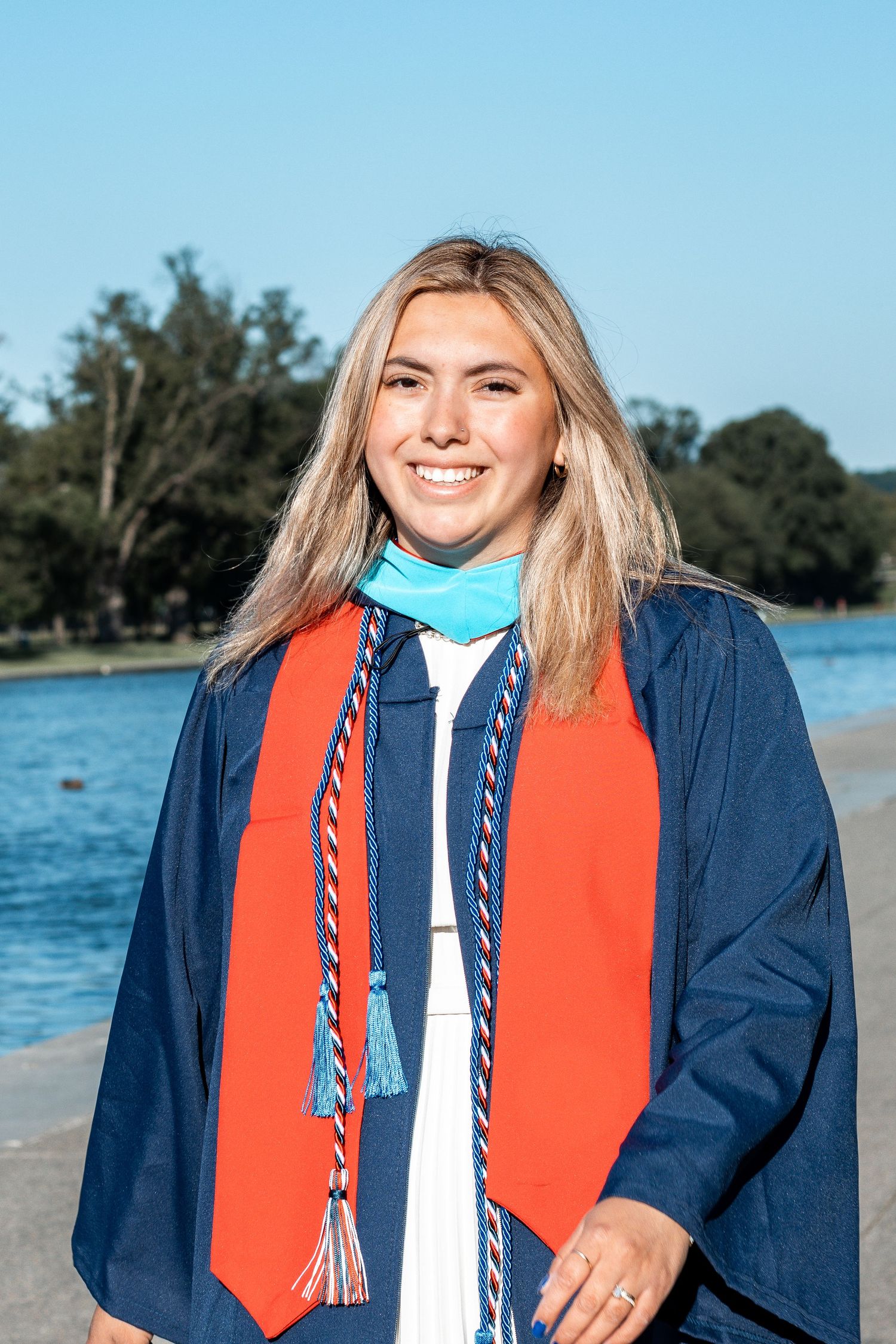 Graduate in navy and orange academic regalia poses outdoors near waterfront.
