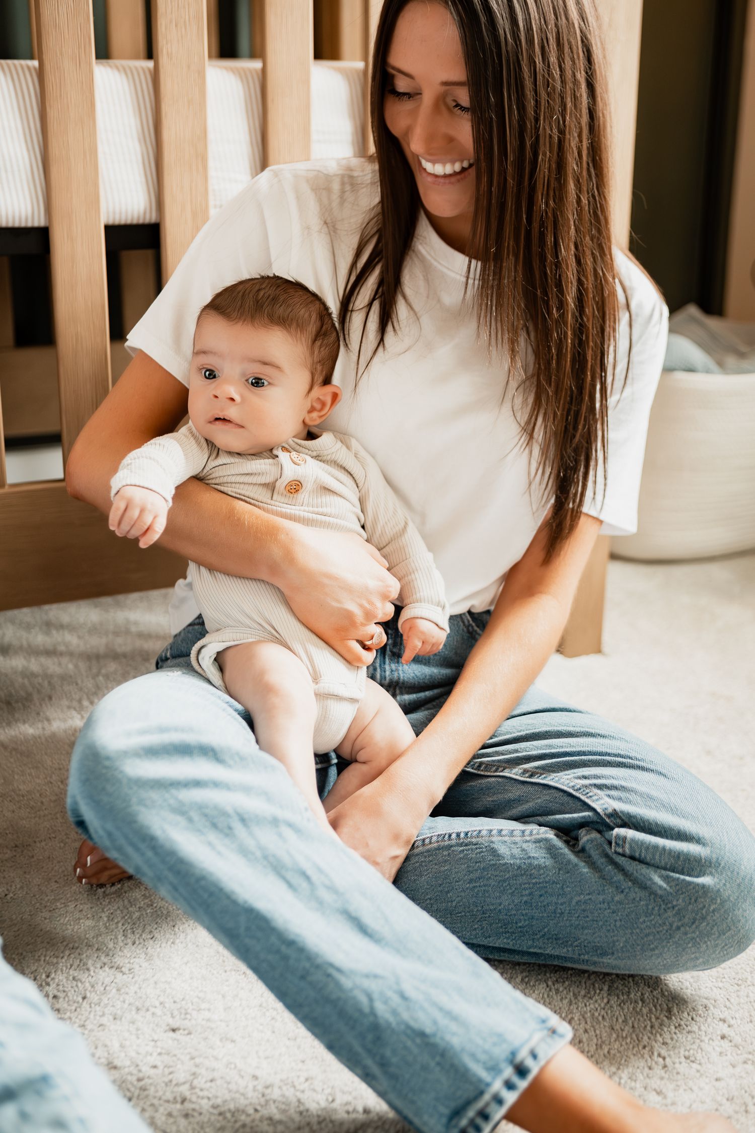 Mother in white shirt and jeans sits on carpet playing with baby wearing a white onesie.