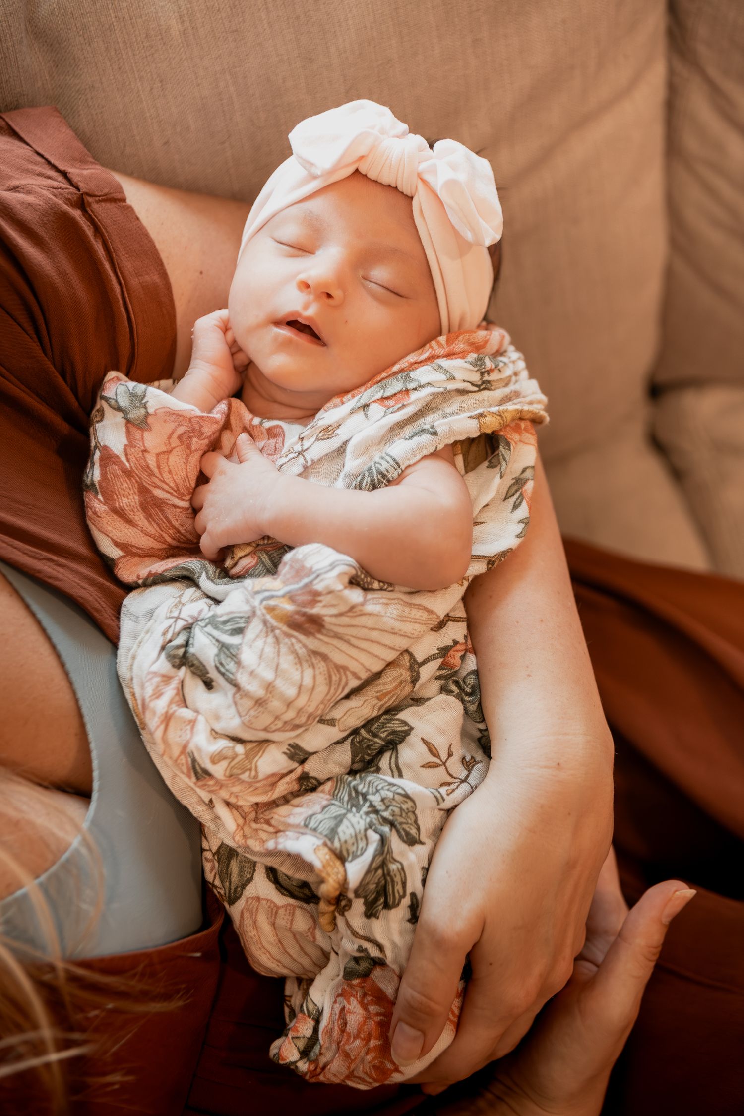 A sleeping newborn baby wearing a white bow headband rests peacefully in floral printed swaddle wrap.
