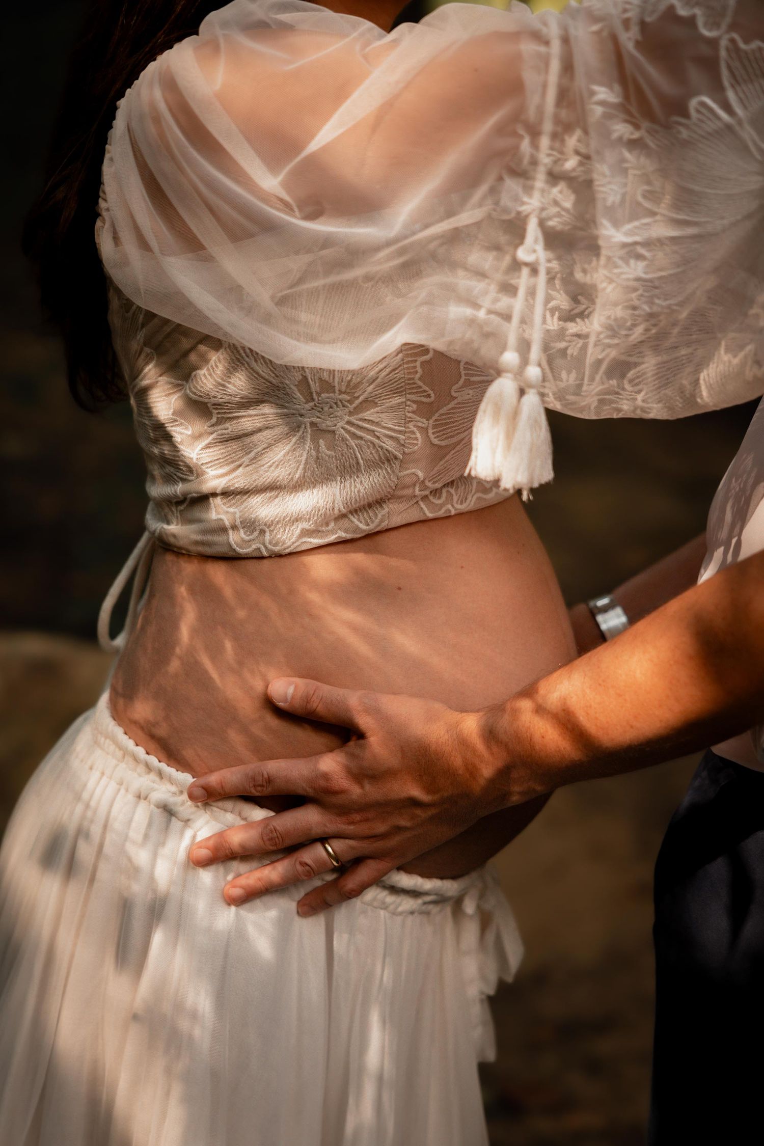 Close-up of a couple's hands on a pregnant woman's belly, wearing wedding rings and elegant white lace attire.