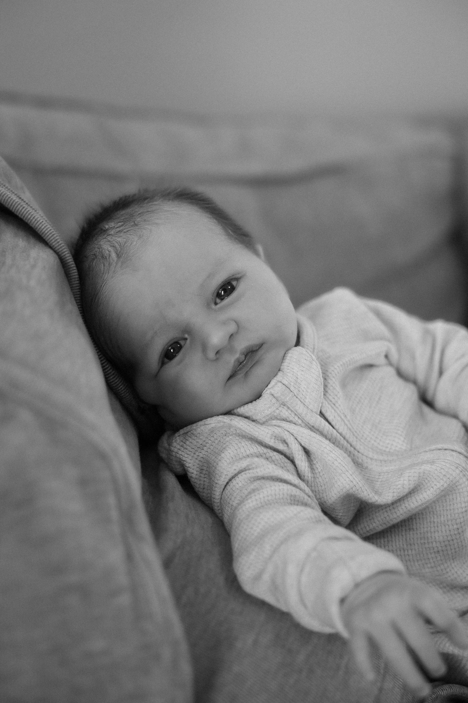 Black and white portrait of a newborn baby lying on a couch cushion, wearing a textured onesie.