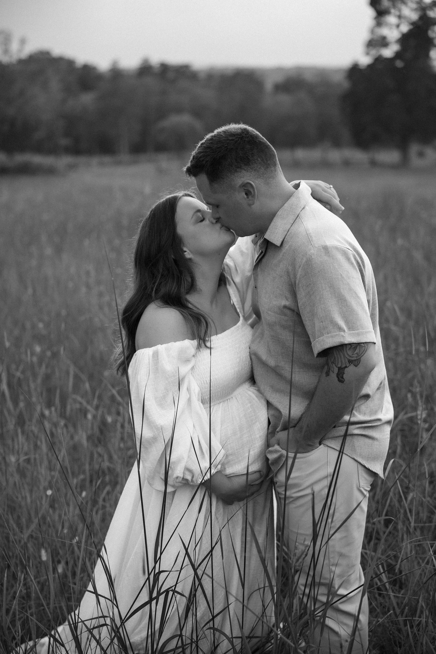 Black and white photo of couple sharing tender kiss in tall grass field during maternity photoshoot.