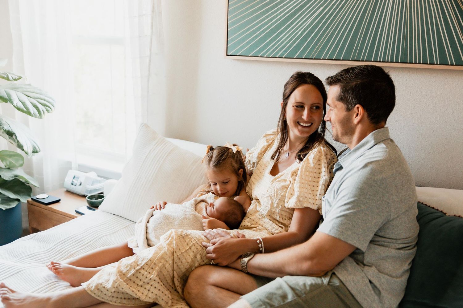 Happy family sitting on a couch, mother holding newborn baby with toddler daughter and smiling father in bright living room.