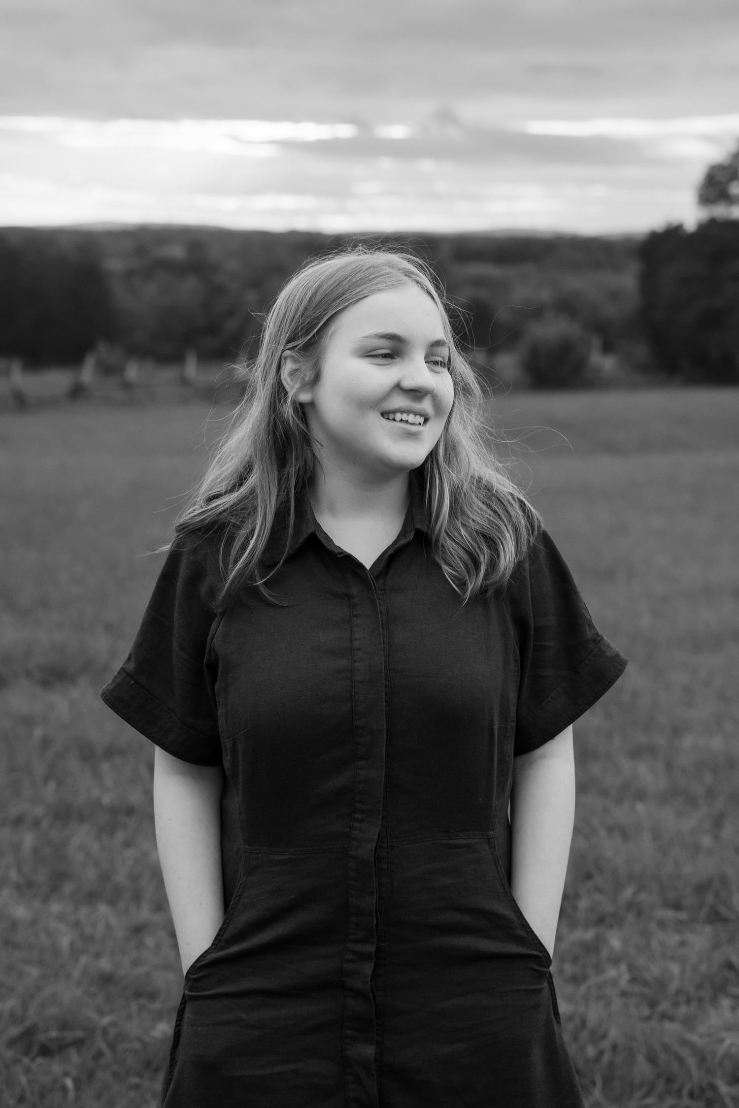 Black and white portrait of a smiling young woman with long hair in a dark dress standing in a rural field.