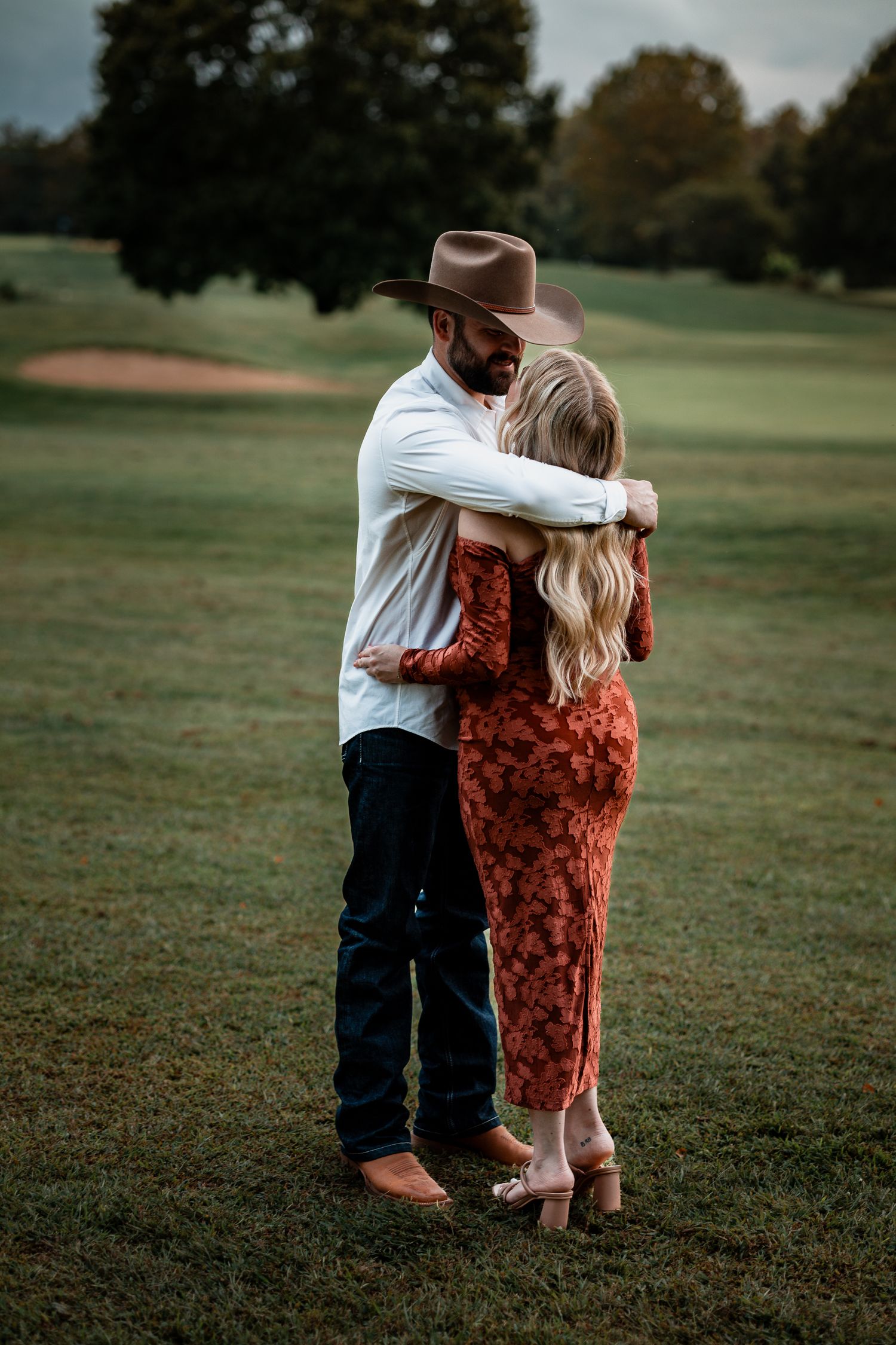 Two people embrace while dancing together on a grassy field at dusk, one in a red dress and one in white.