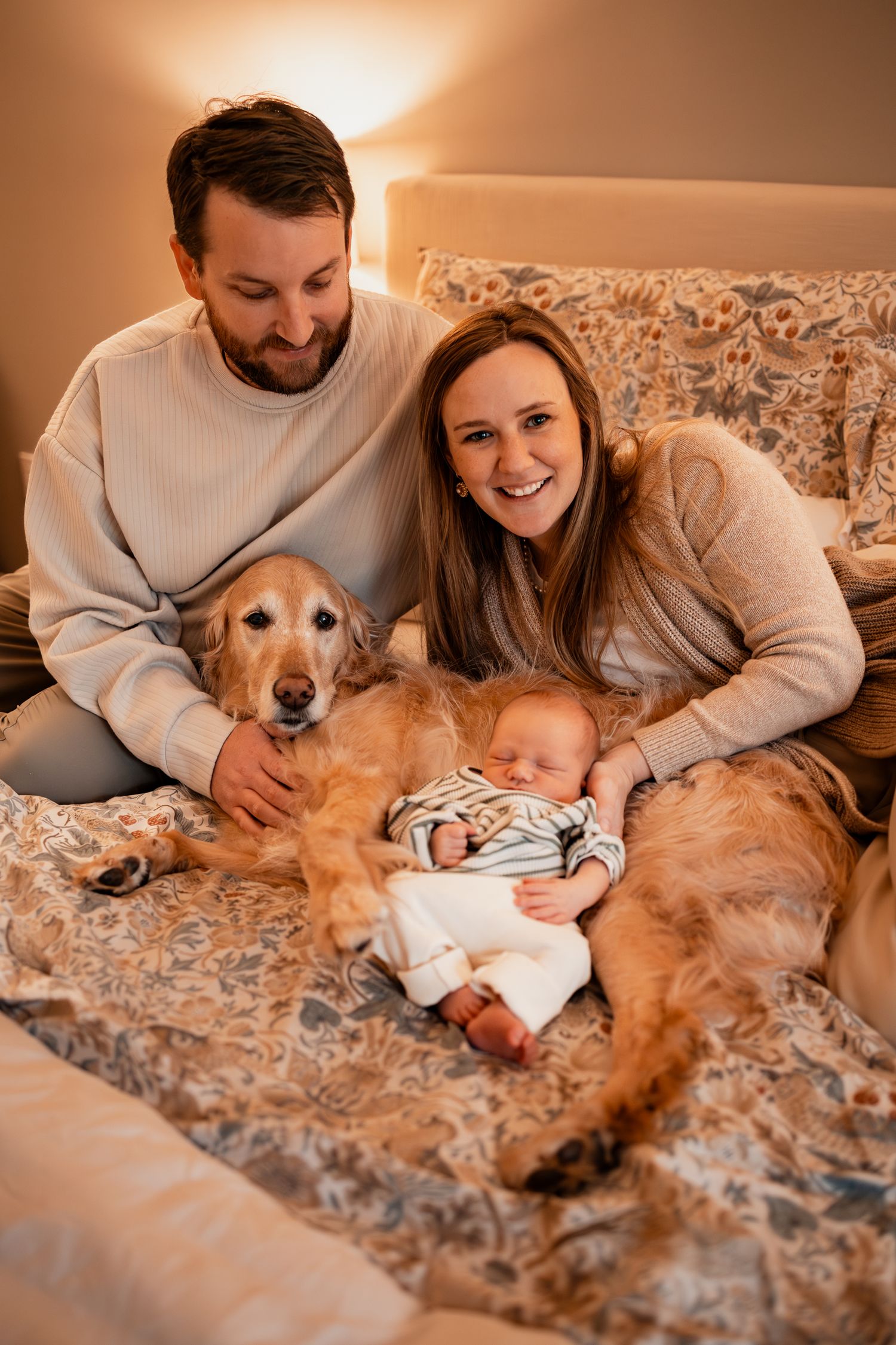 Family lounging on bed with golden retriever and sleeping newborn.
