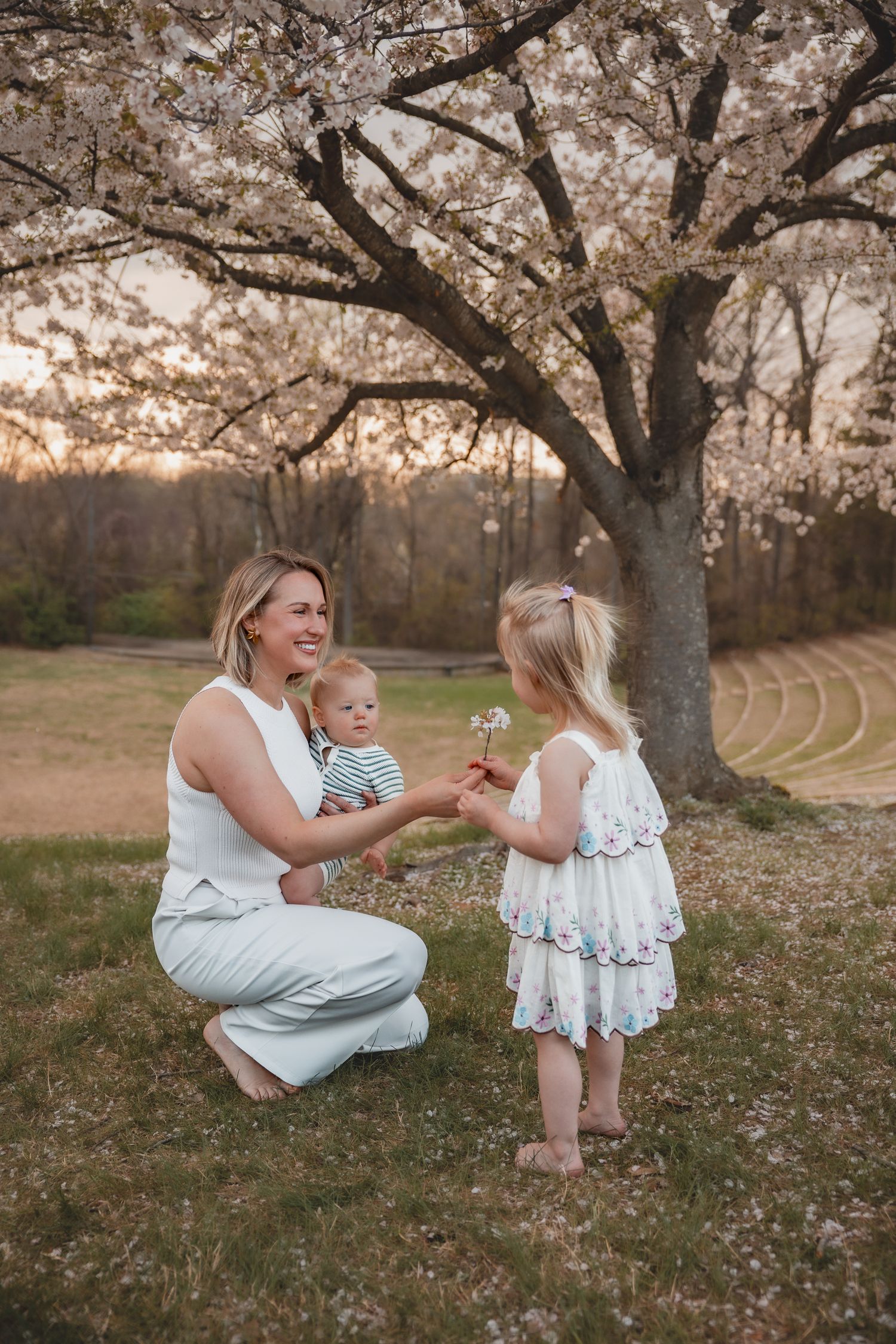 Mother crouching with baby holds dandelion with toddler girl in white dress under blooming cherry tree at sunset.