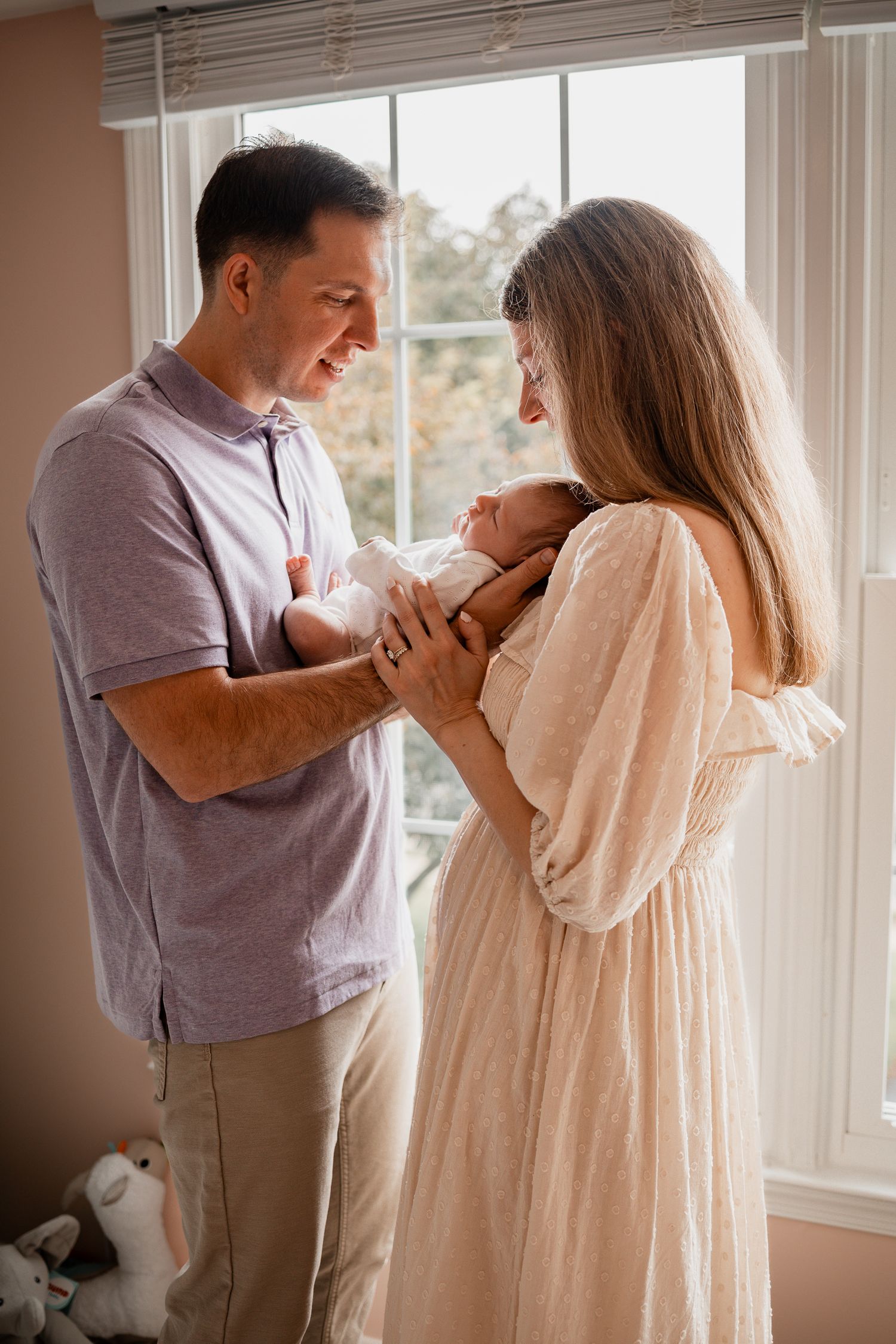 A couple smiles lovingly at their newborn baby by a bright window in a cozy home nursery setting.