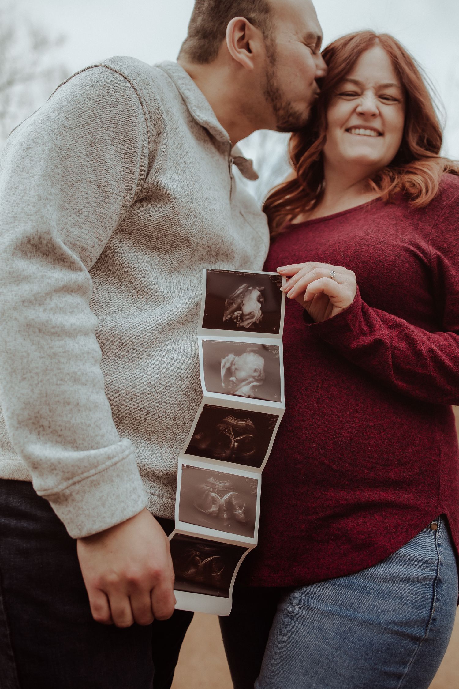 A couple in burgundy and gray hold ultrasound photos while sharing an intimate moment together outdoors.