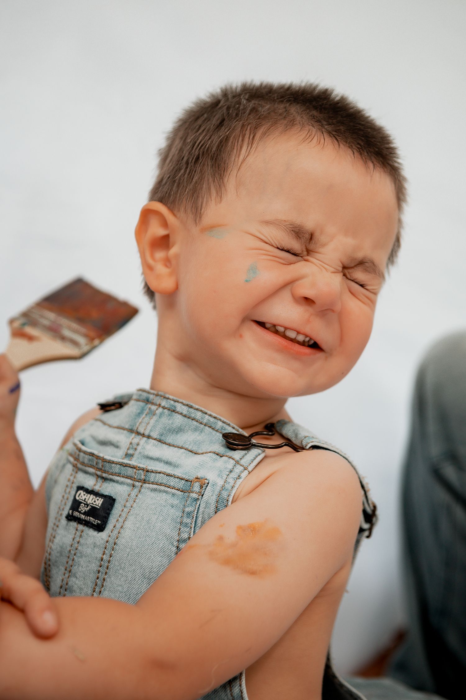 Child in denim overalls scrunches face and squints eyes with a paint brush touching their arm.
