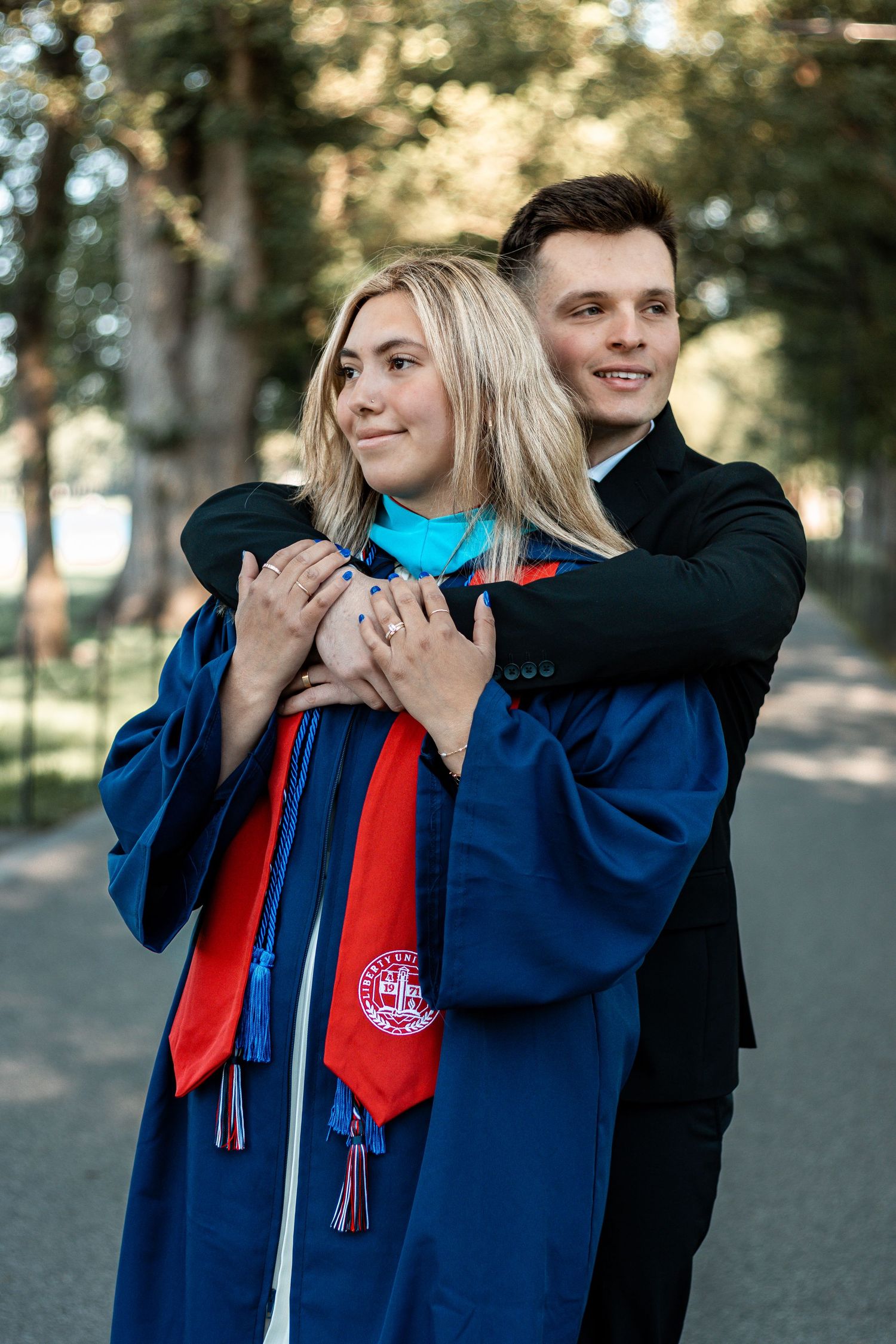 Two graduates share a celebratory moment in front of trees wearing blue and red academic regalia.