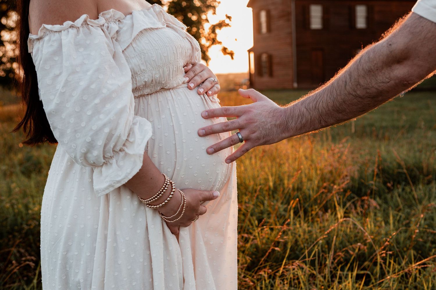 Gentle hand touches pregnant belly in white dress at sunset in grassy field.