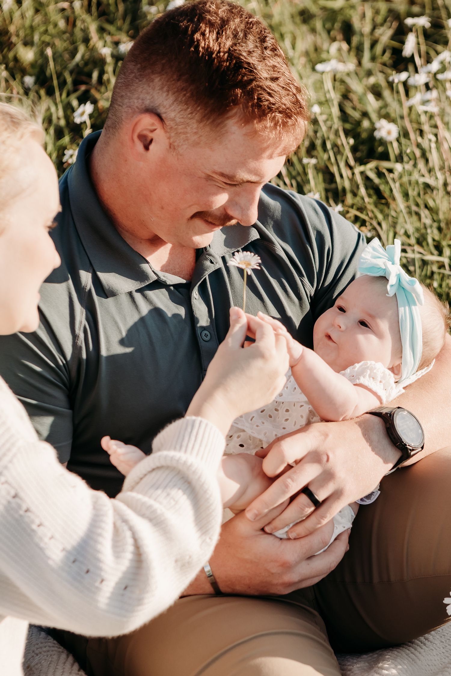 Sweet outdoor family moment with parent holding infant in polka dot dress and floral headband in sunny meadow with wildflowers.