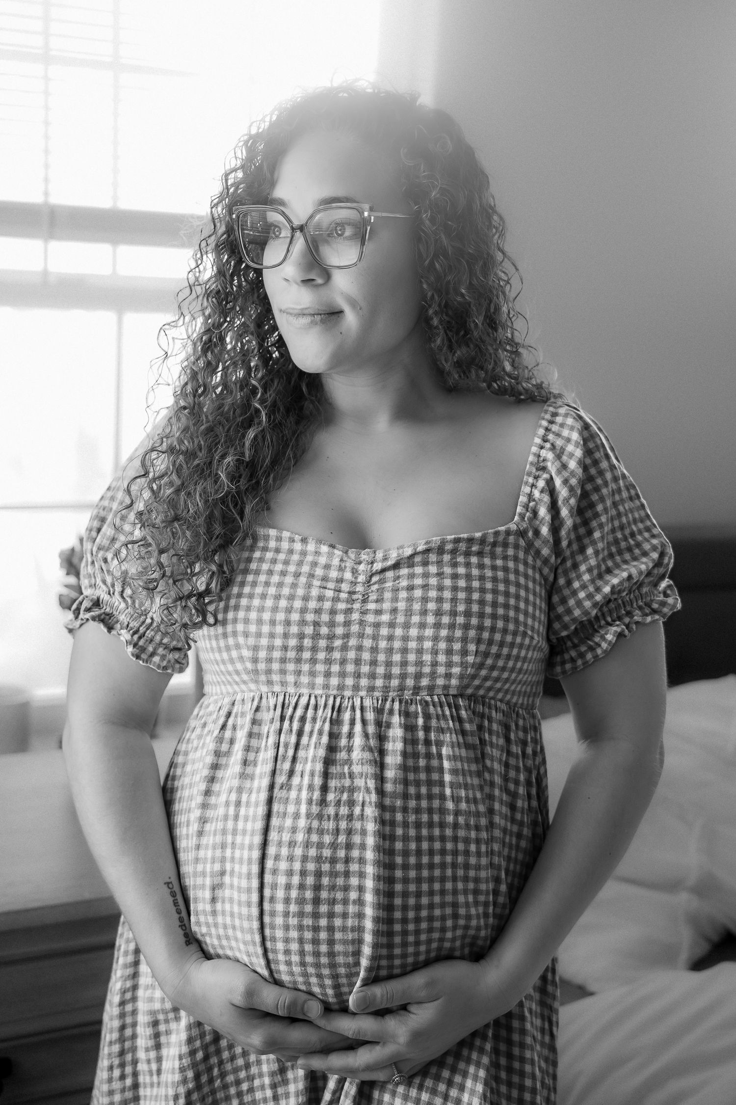 Black and white photo of a pregnant woman with curly hair and glasses, wearing a plaid dress, cradling her baby bump.