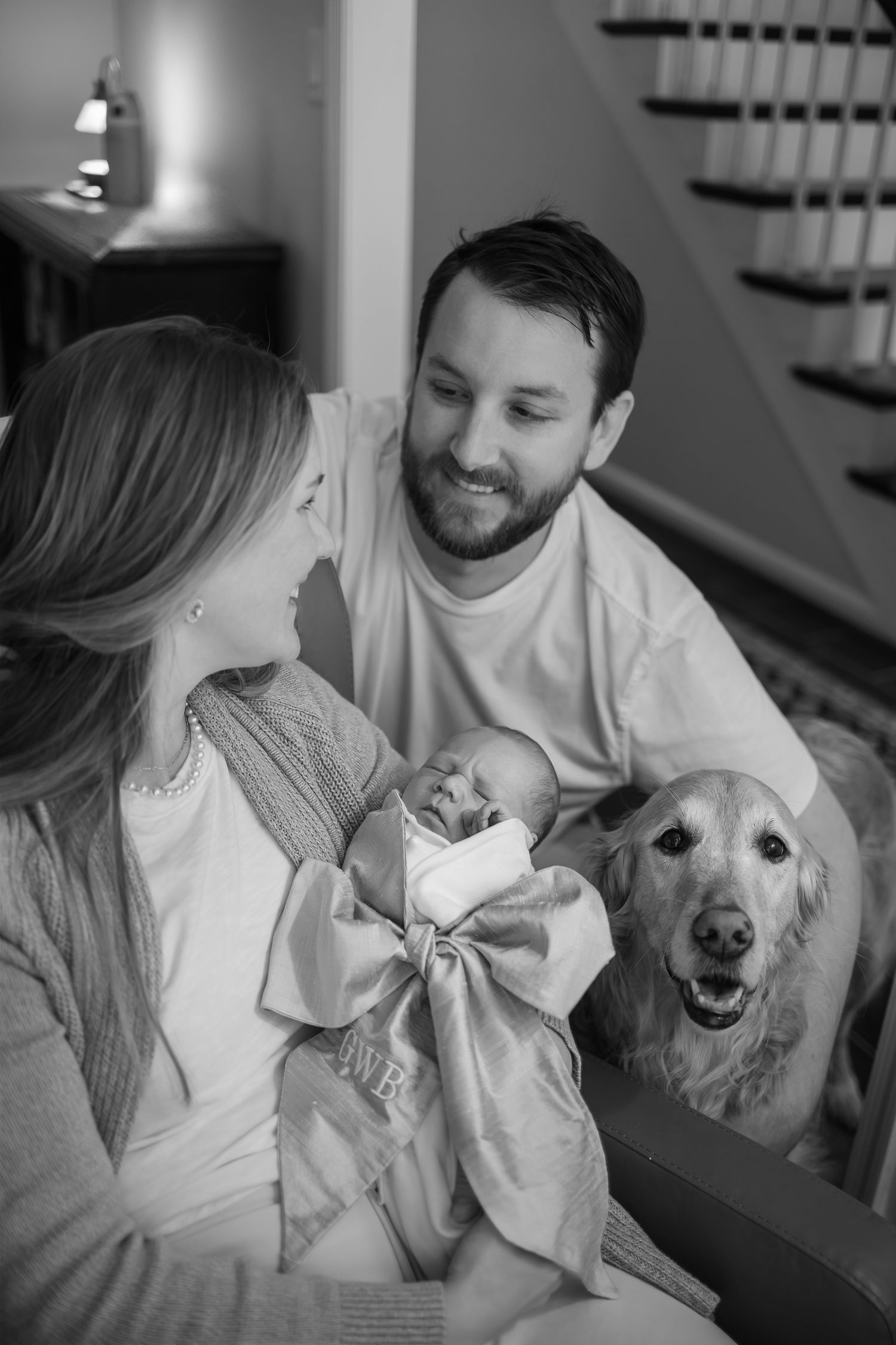 A newborn baby is cradled between parents with their golden retriever dog looking on in a black and white photo.