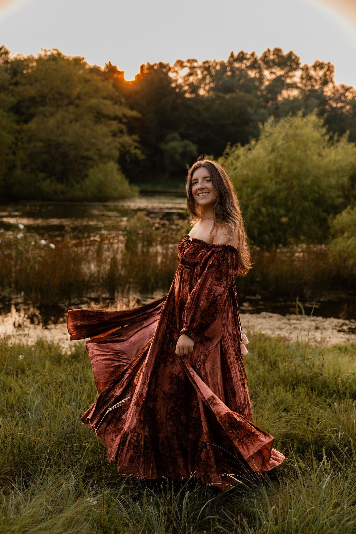 Woman in flowing rust velvet off-shoulder gown twirling at golden hour near a serene pond surrounded by lush greenery.