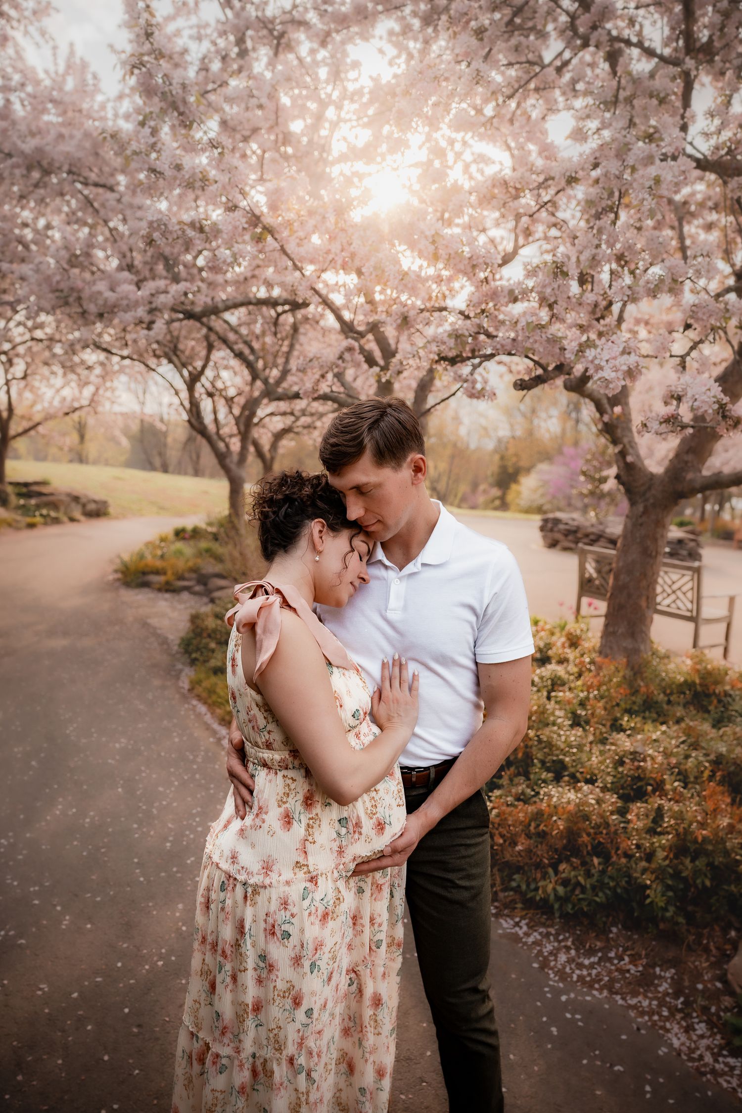A couple embraces tenderly under blooming cherry blossom trees in a romantic spring park setting.