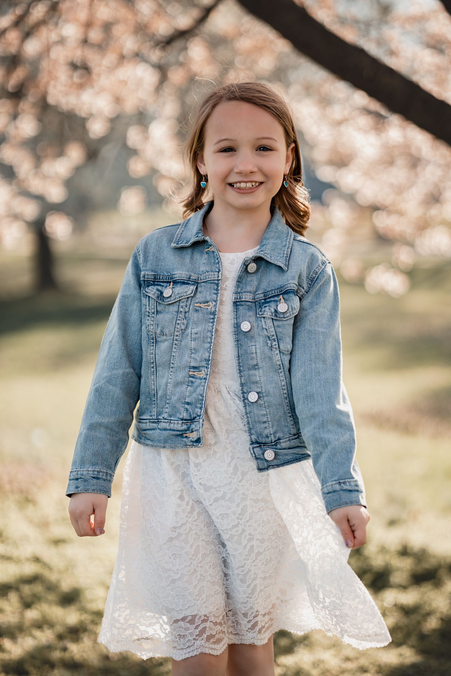 A smiling young girl in a white lace dress and denim jacket poses outdoors near blooming cherry blossom trees.