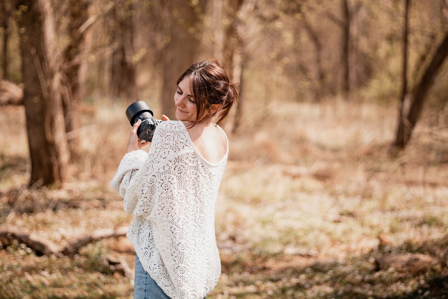 Woman in white lace sweater holding camera in forest, smiling over shoulder, outdoor photography portrait.