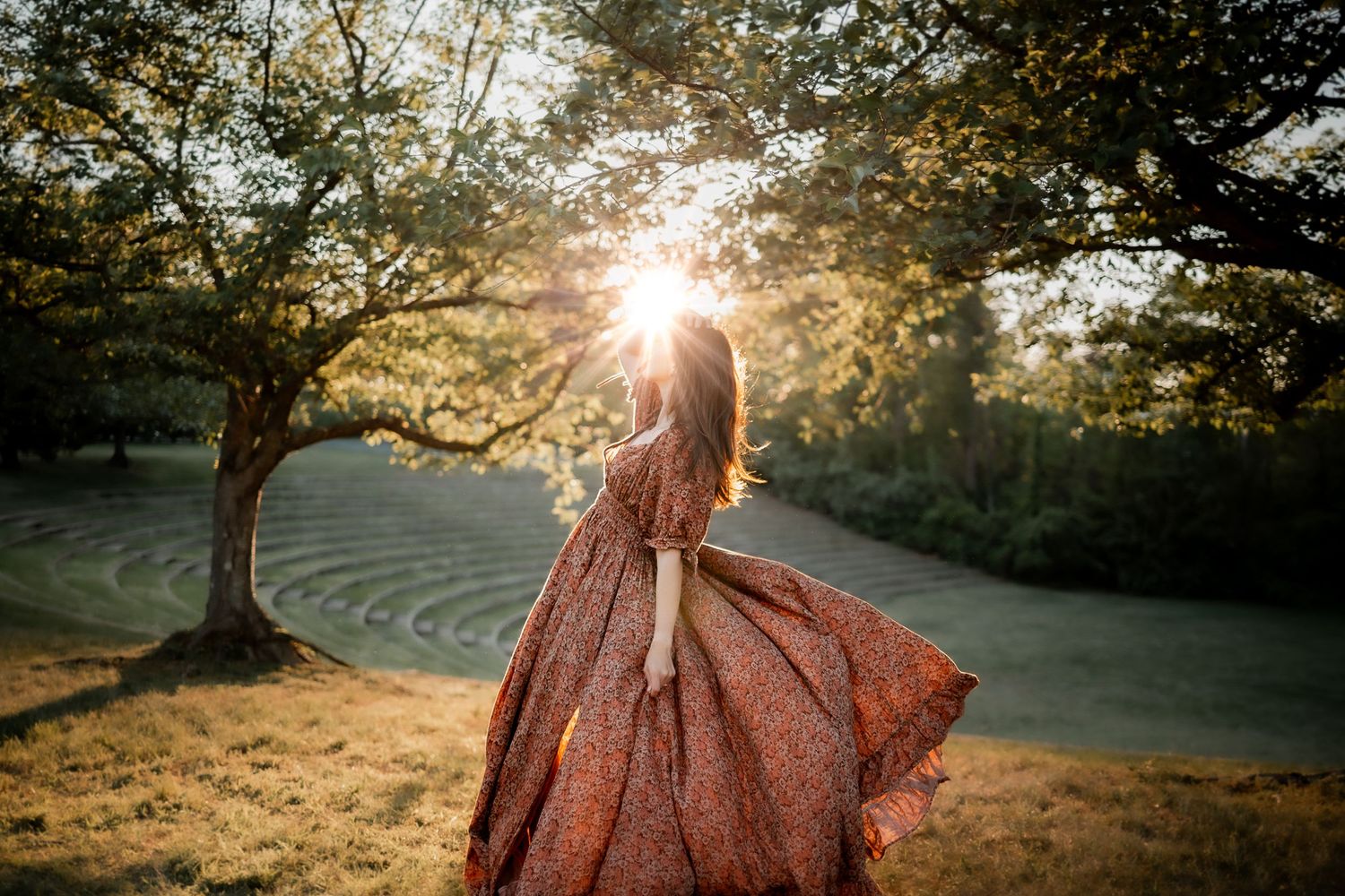 Woman in flowing red floral gown twirling in sunlit park at golden hour with trees and manicured lawn.