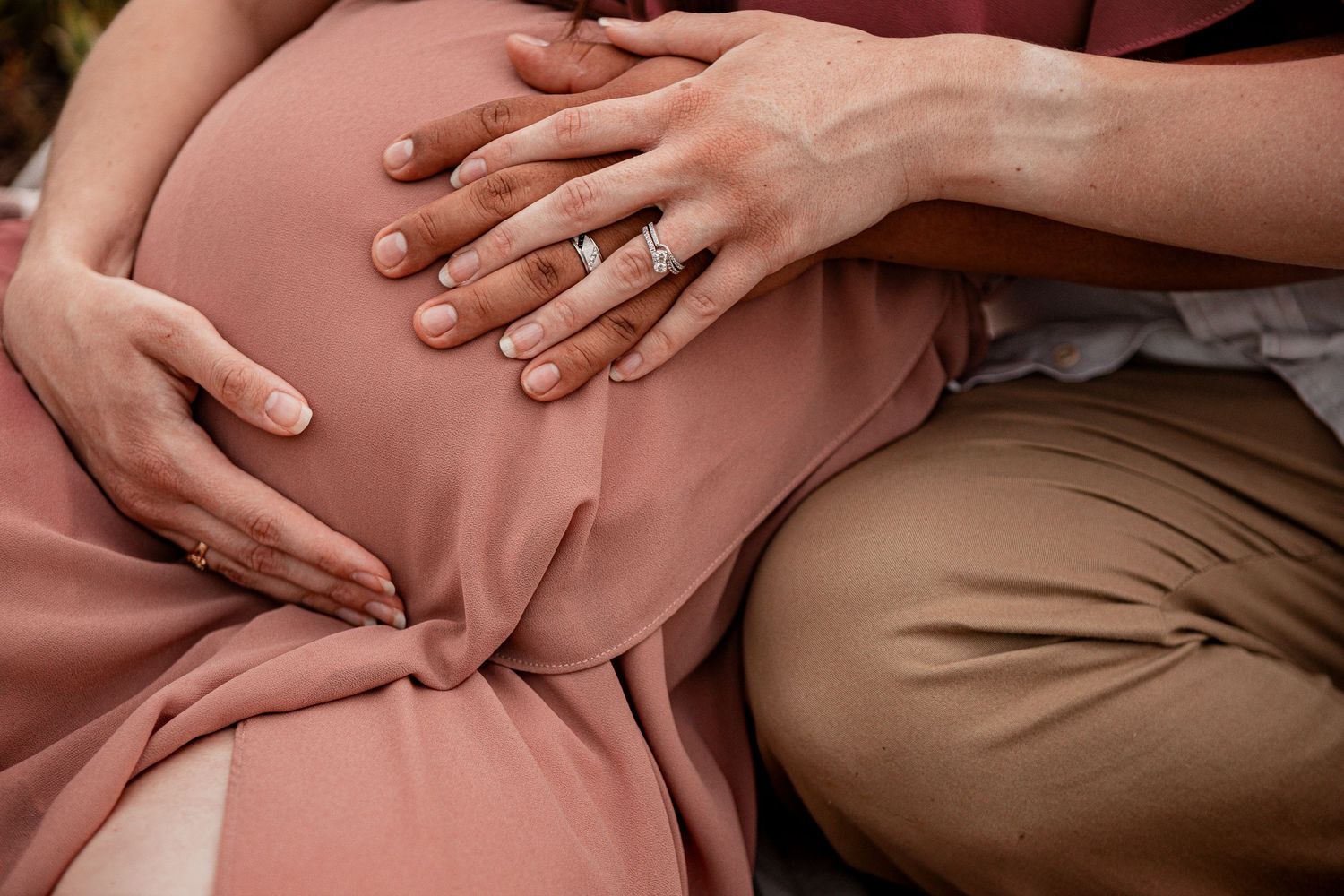 Hands tenderly rest on a pregnant belly covered in a soft pink dress, with a wedding ring visible.