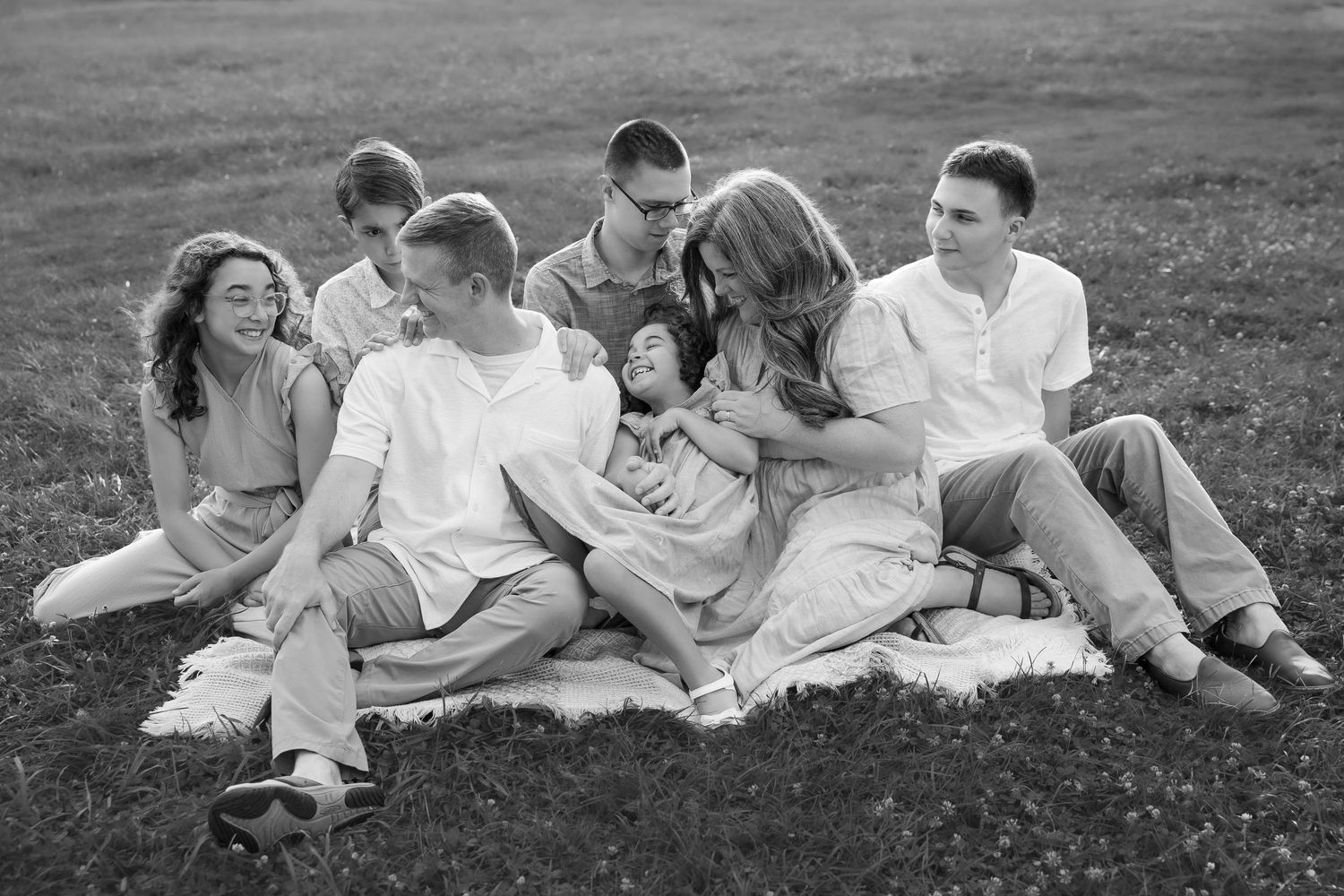 Black and white family photo of seven people sitting together on a blanket in a grassy field, laughing and embracing.