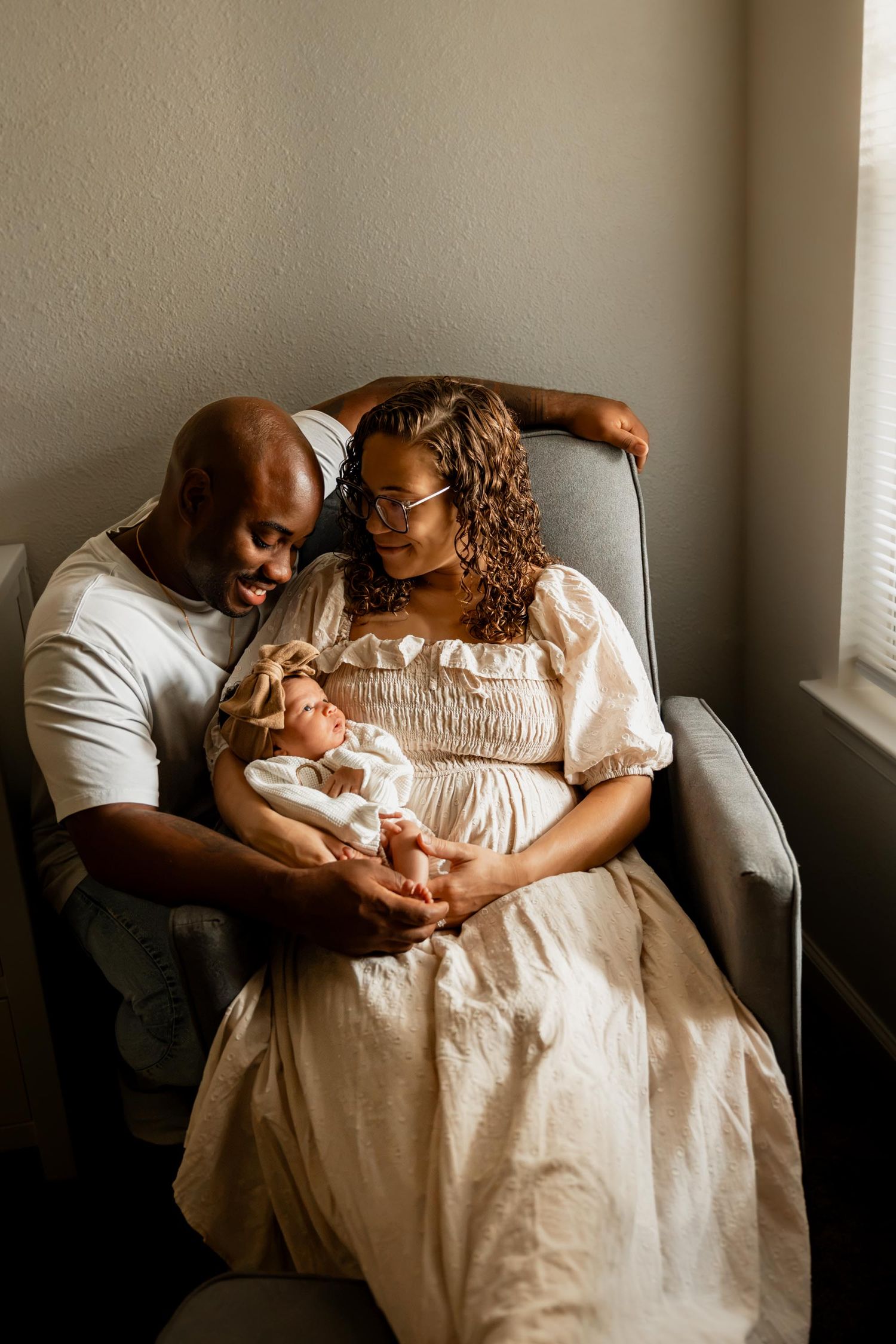 A couple gazes lovingly at their newborn baby, seated by a window in a cozy gray chair.