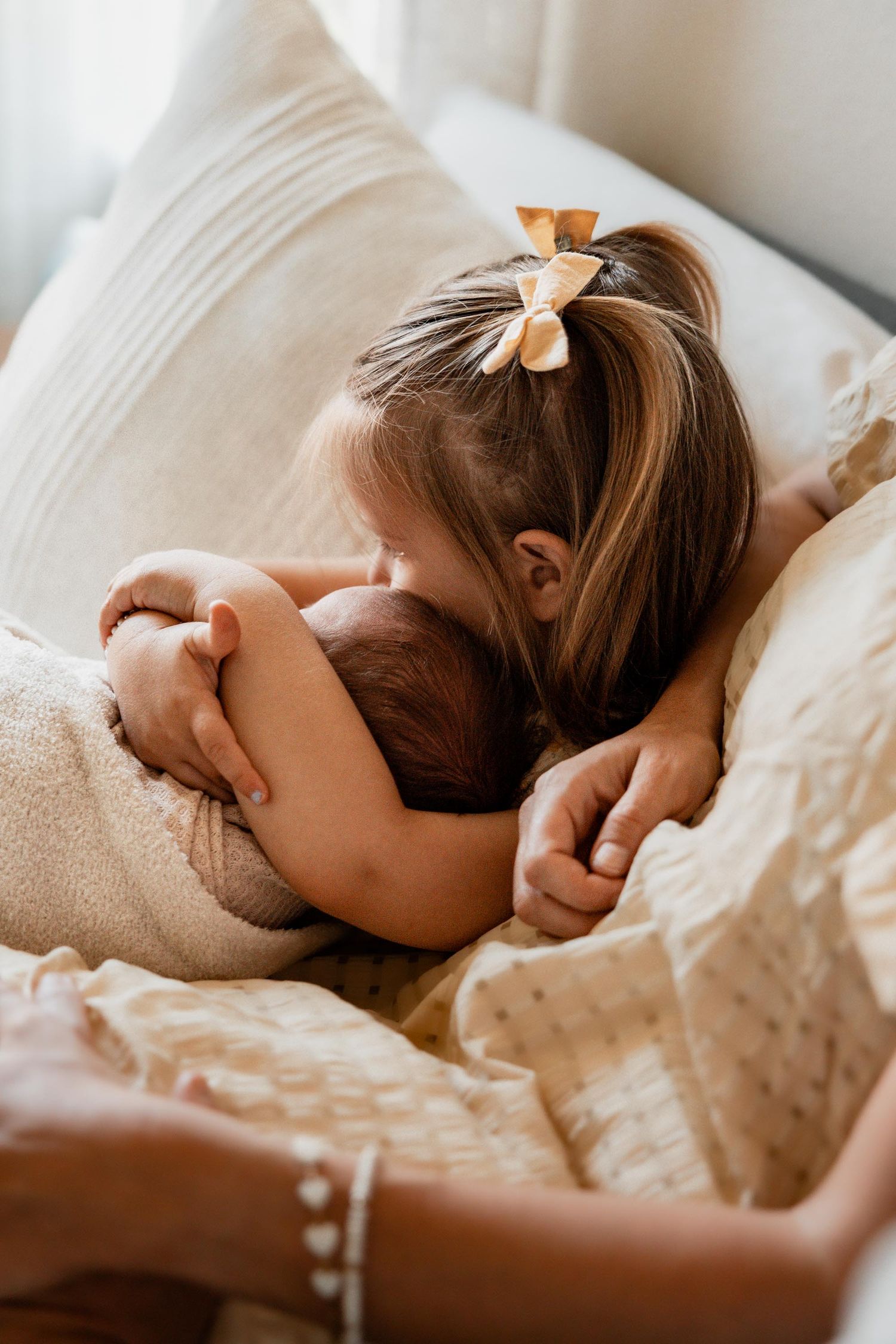 Young girl with bow gently hugging newborn baby on cozy cream bedding.