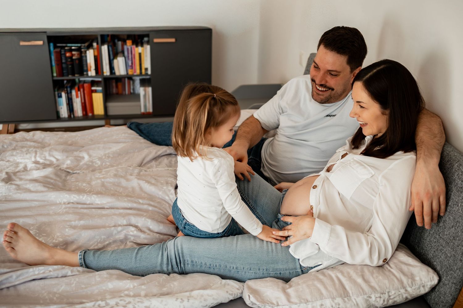 Family relaxing together on white bed while looking at mobile devices and tablets.