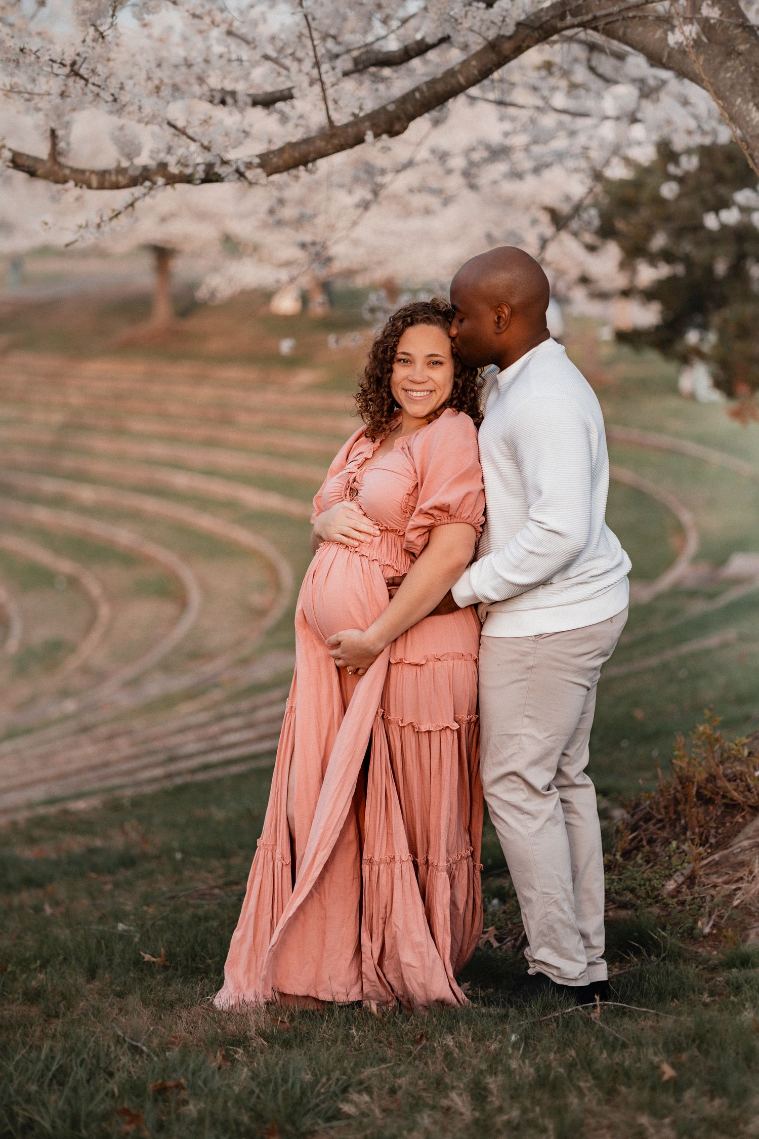 Pregnant couple embracing under cherry blossom tree, woman in pink maxi dress, man in white shirt, golden hour maternity photo.