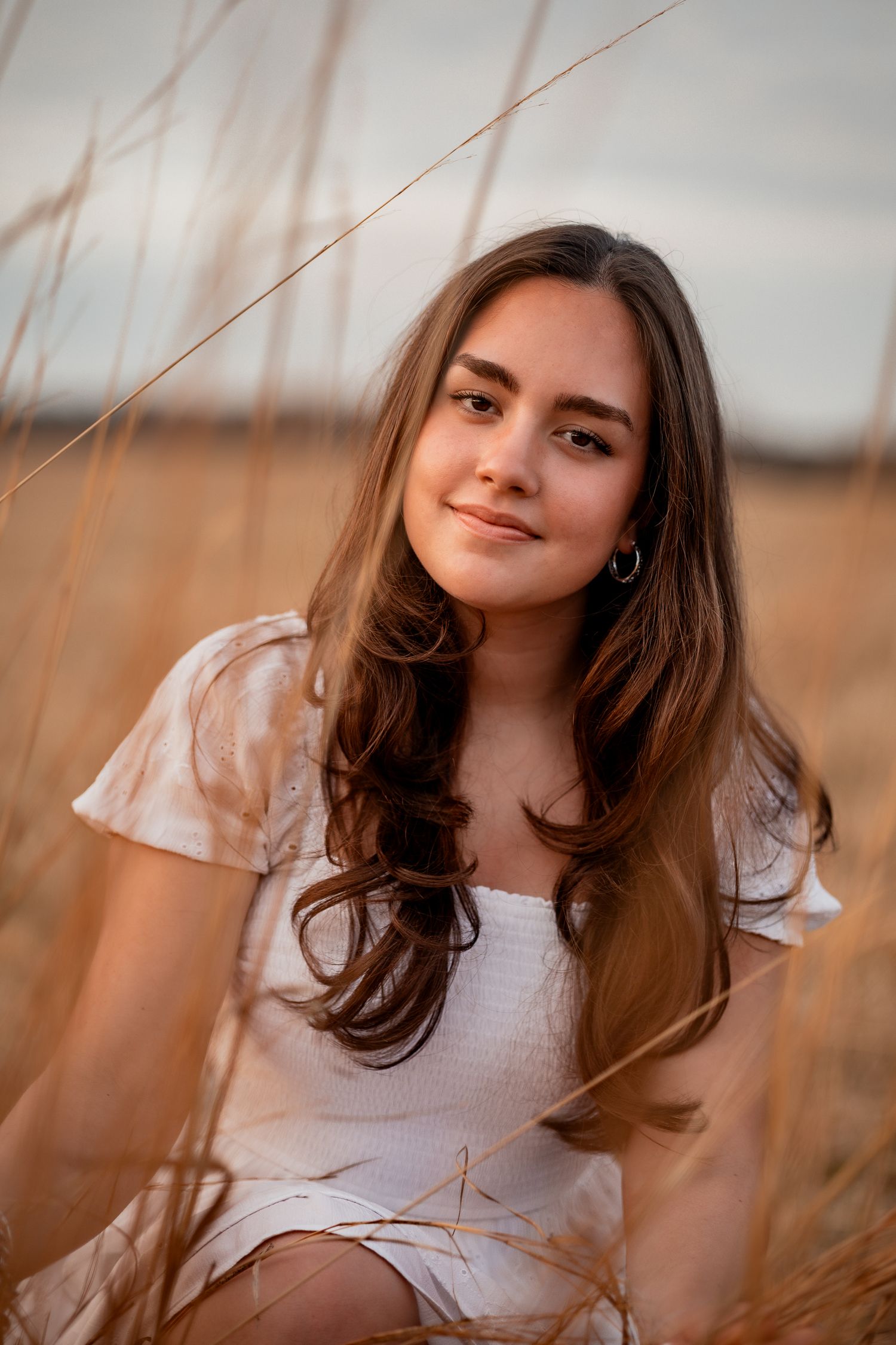 Young woman with long brown hair in white dress sitting in golden field at sunset.