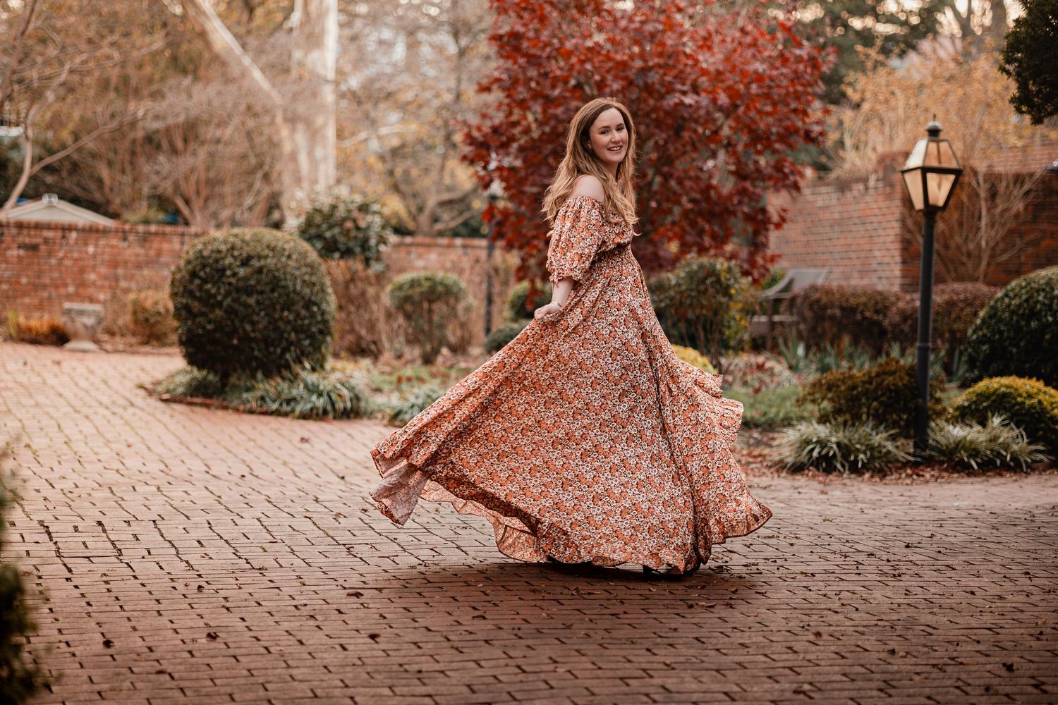 A person in a flowing rust-colored dress twirls gracefully in a garden setting during autumn.