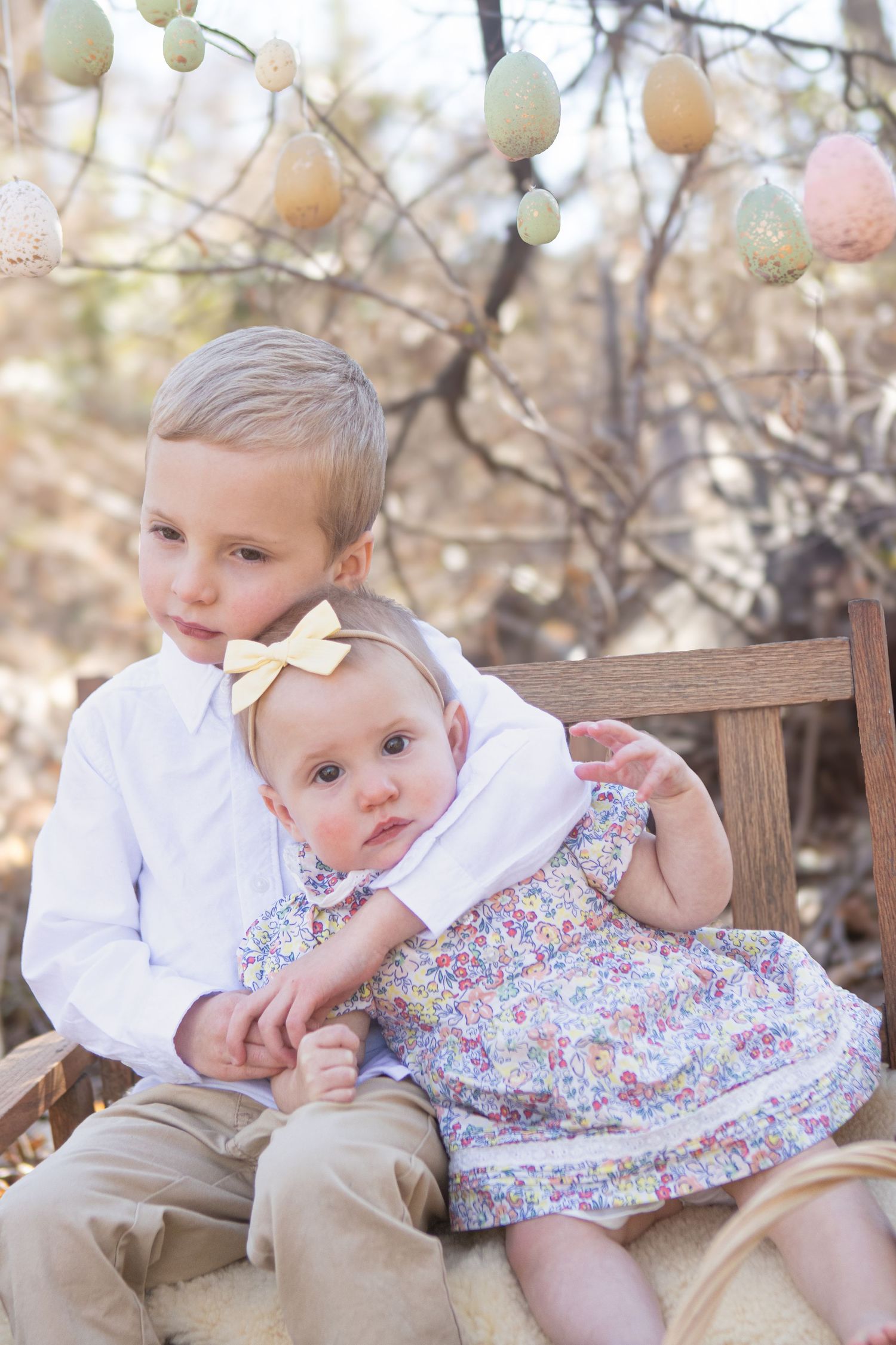 Children sitting on bench with pastel Easter eggs hanging from tree branches in background.