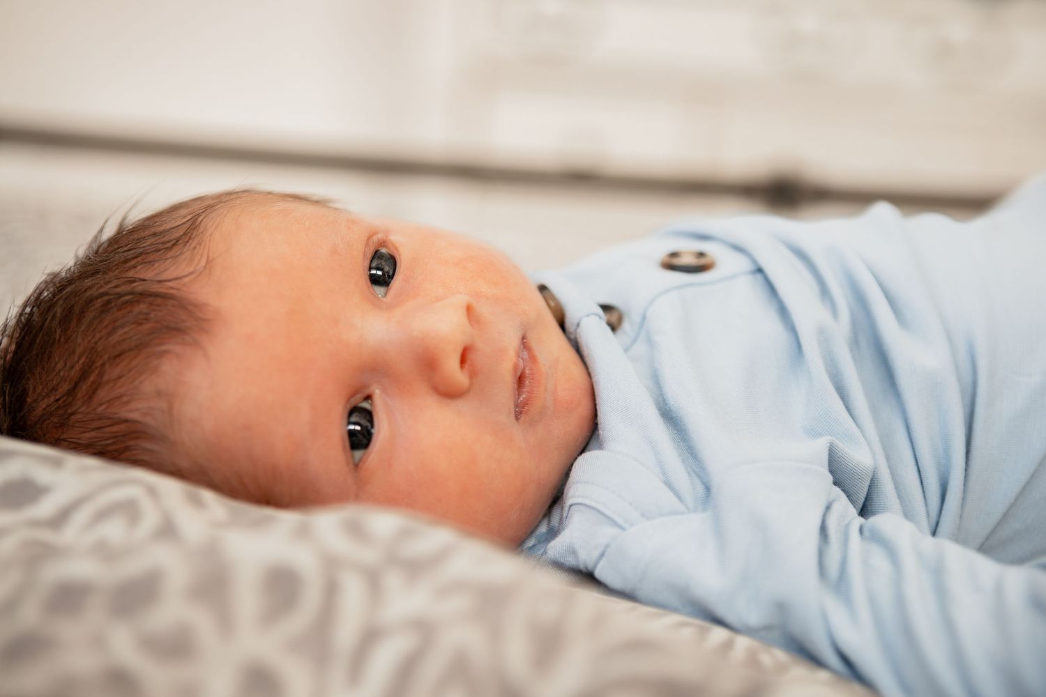 Cute newborn baby wearing light blue onesie looking up while resting against white bedding.