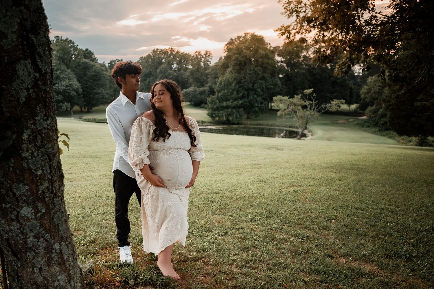 A couple in light summer attire share an intimate moment under a tree during golden hour in a lush park setting.