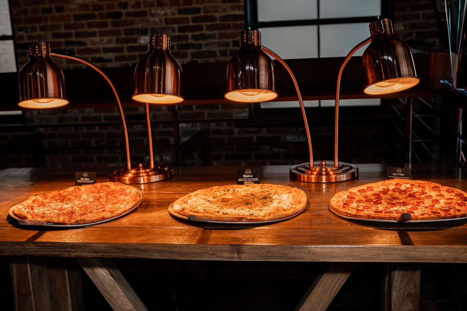 Row of illuminated pendant lamps hanging above pizzas on wooden tables in restaurant setting.